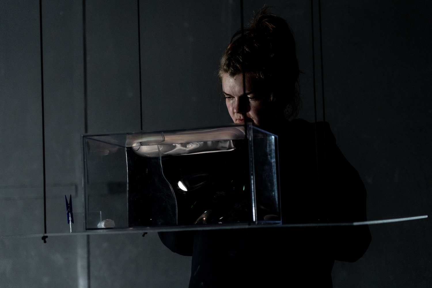 Schauspielerin schaut im Bühnendunkel auf einen aquariumähnlichen Kasten, in dem eine Videoprojektion angedeutet ist.