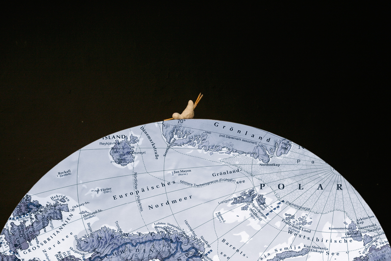 Im unteren Bildteil ist eine zur Hälfte angeschnittene Weltkugel aus Atlaspapier. Oben liegt mittig eine kleine, nicht genau erkennbare Figur, die vielleicht aus Ton ist. Der Rest des Bildes ist schwarz.
