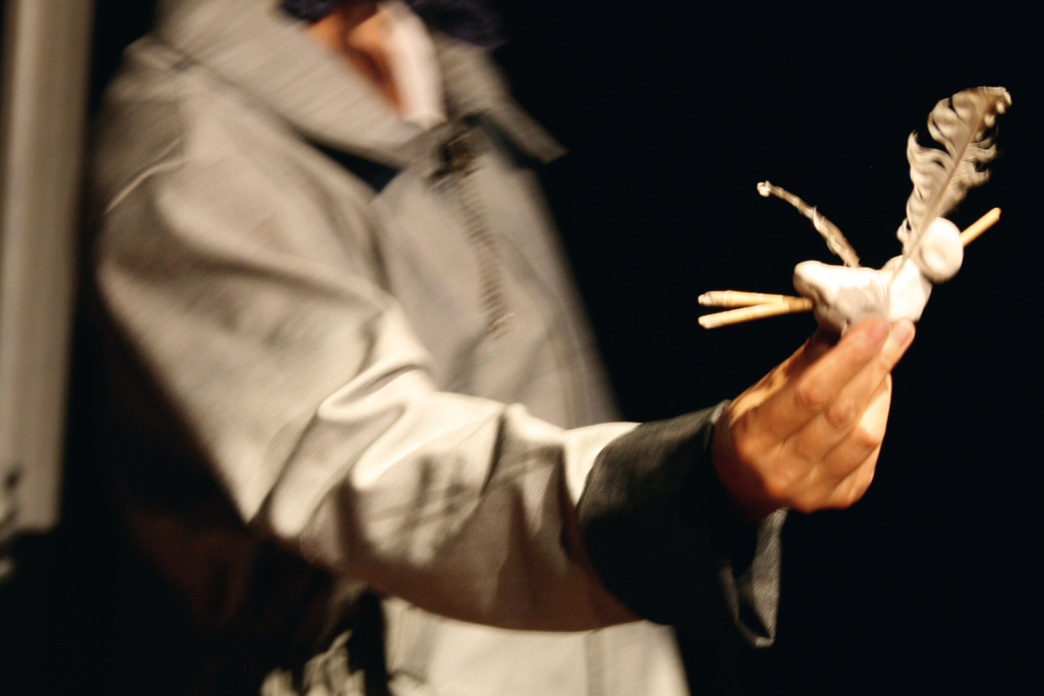 In Nahaufnahme ist schemenhaft der Oberkörper und Arm einer Person auf dem Bühnenraum zu sehen. In der Hand hält sie eine kleine Figur aus weißer Knete mit Stöckchen als Armen und einer Feder.