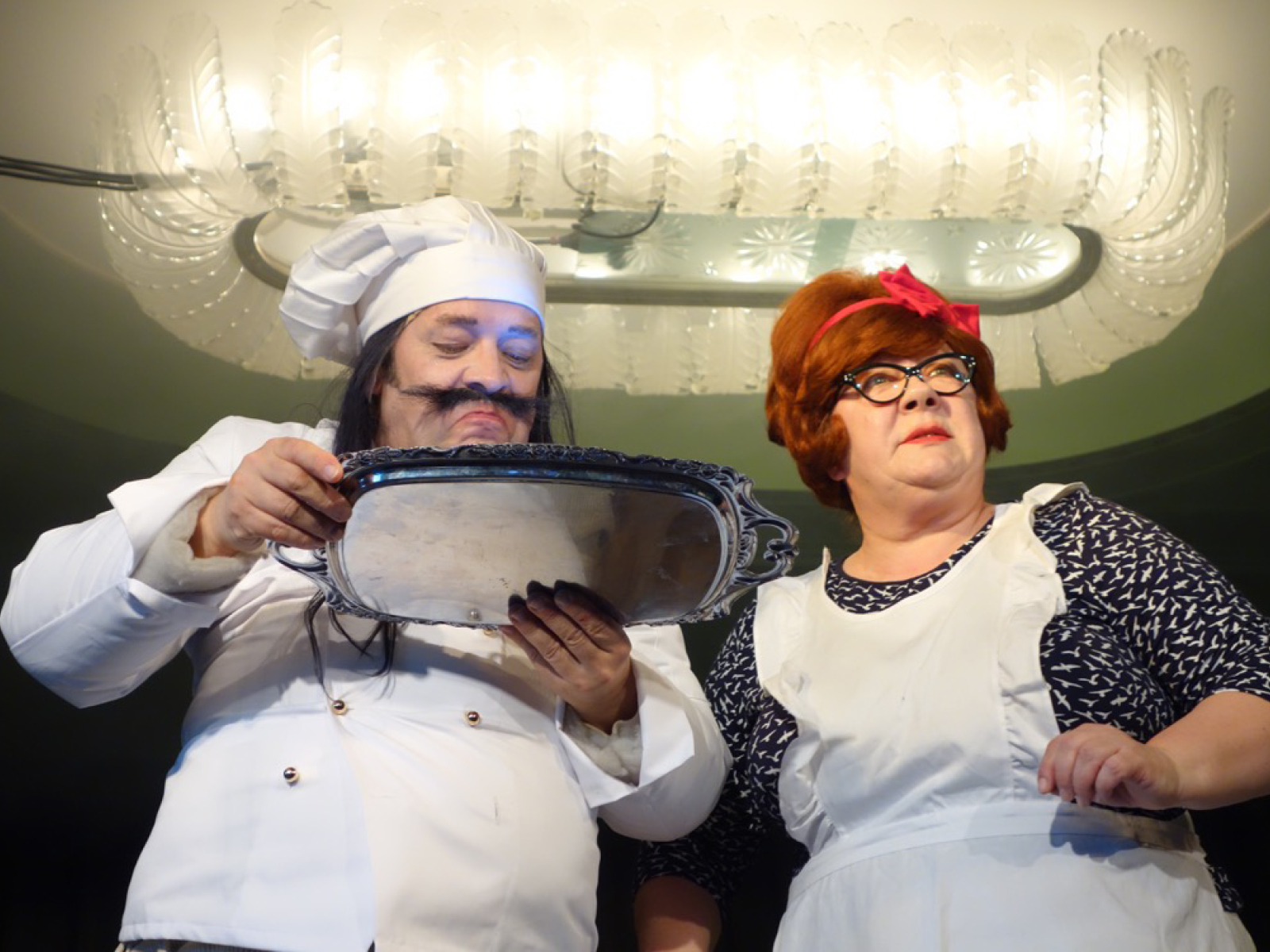 Koch mit Kochmütze und Schnurrbart schaut auf eine Silberplatte. Neben ihm eine rothaarige Frau mit Brille und weißer Schürze. Beide sind etwa 50 Jahre alt.
