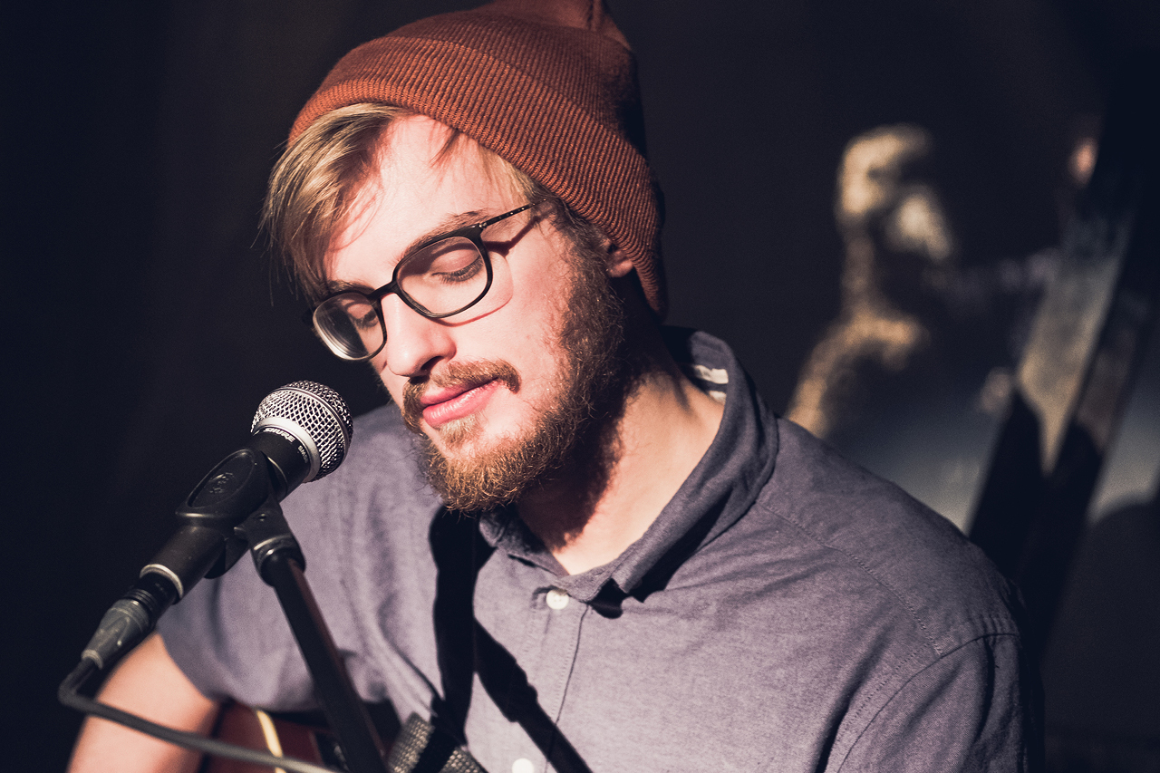 Ein Musiker mit Bart, Brille und roter Mütze in Nahaufnahme vor einem Mikrofon sitzend. Nur sein Oberkörper ist sichtbar. Im Hintergrund ist schemenhaft eine Gänse-Puppe erkennbar.