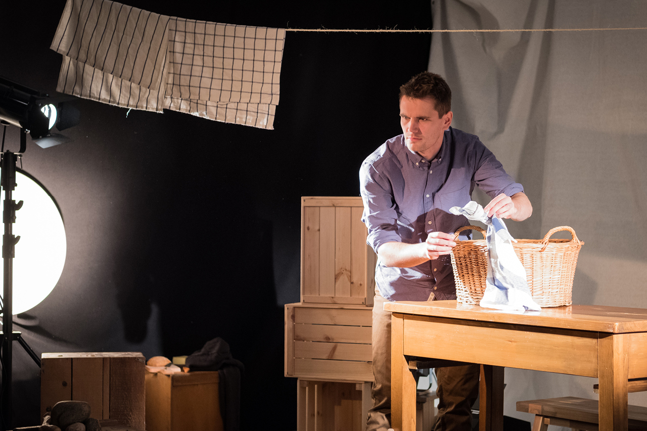 Ein Schauspieler hantiert mit einem geknoteten Geschirrhandtuch auf der Bühne. Er steht an einem Tisch, auf dem sich ein Wäschekorb befindet. Über ihm hängt eine Leine mit weiteren Geschirrtüchern. Links außen gibt es eine helle, runde Lichtquelle im Bild.