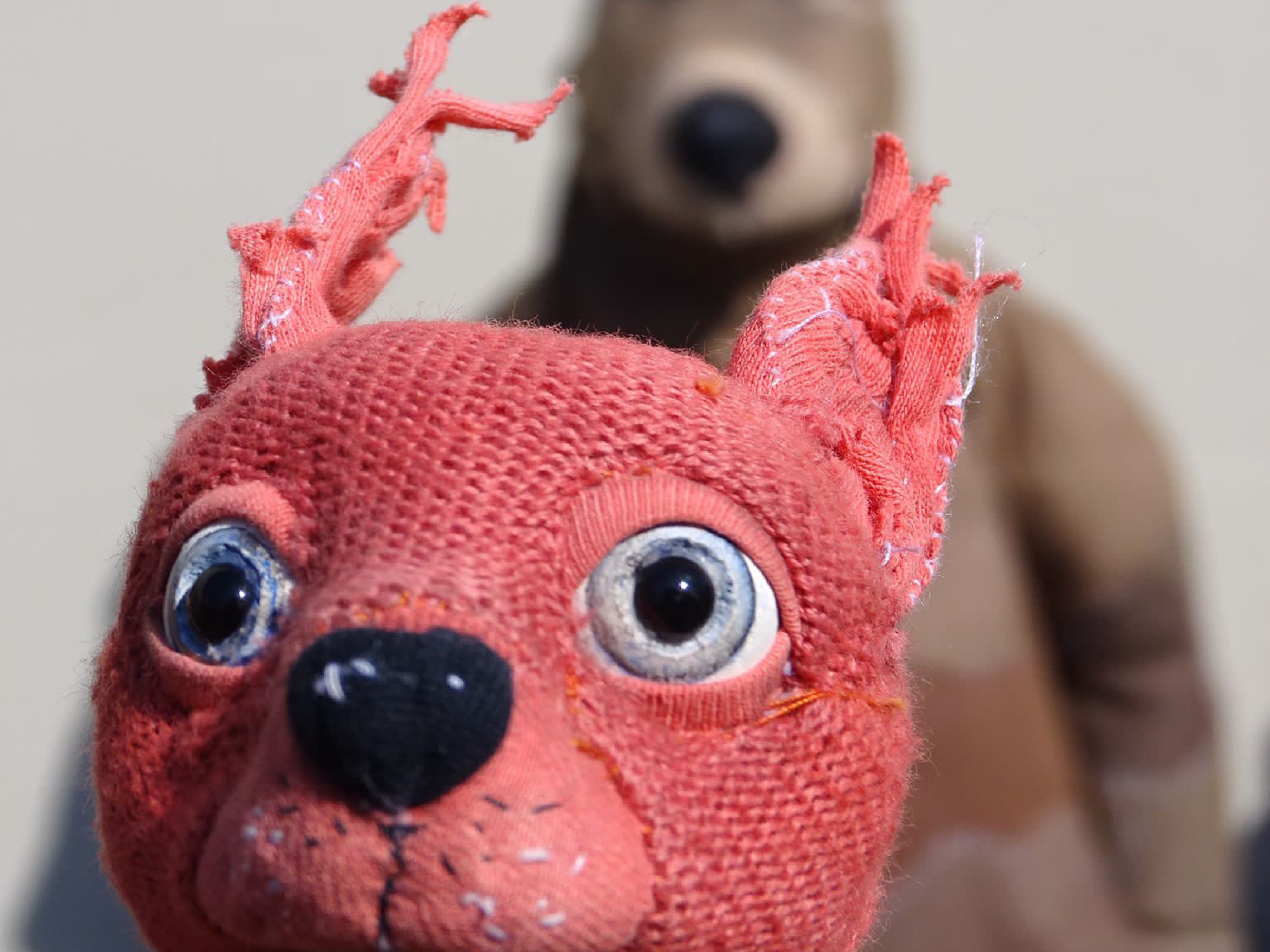 The puppet of a stuffed squirrel stands close to the camera lens. The squirrel has blue eyes and embroidered whiskers. The large puppet of a bear stands in the background.