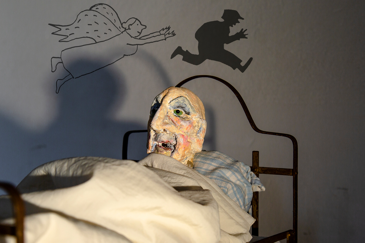 Puppe eines alten Mannes liegt im Bett. Darüber sind Zeichnungen einer Kinderfigur und eines Engels.