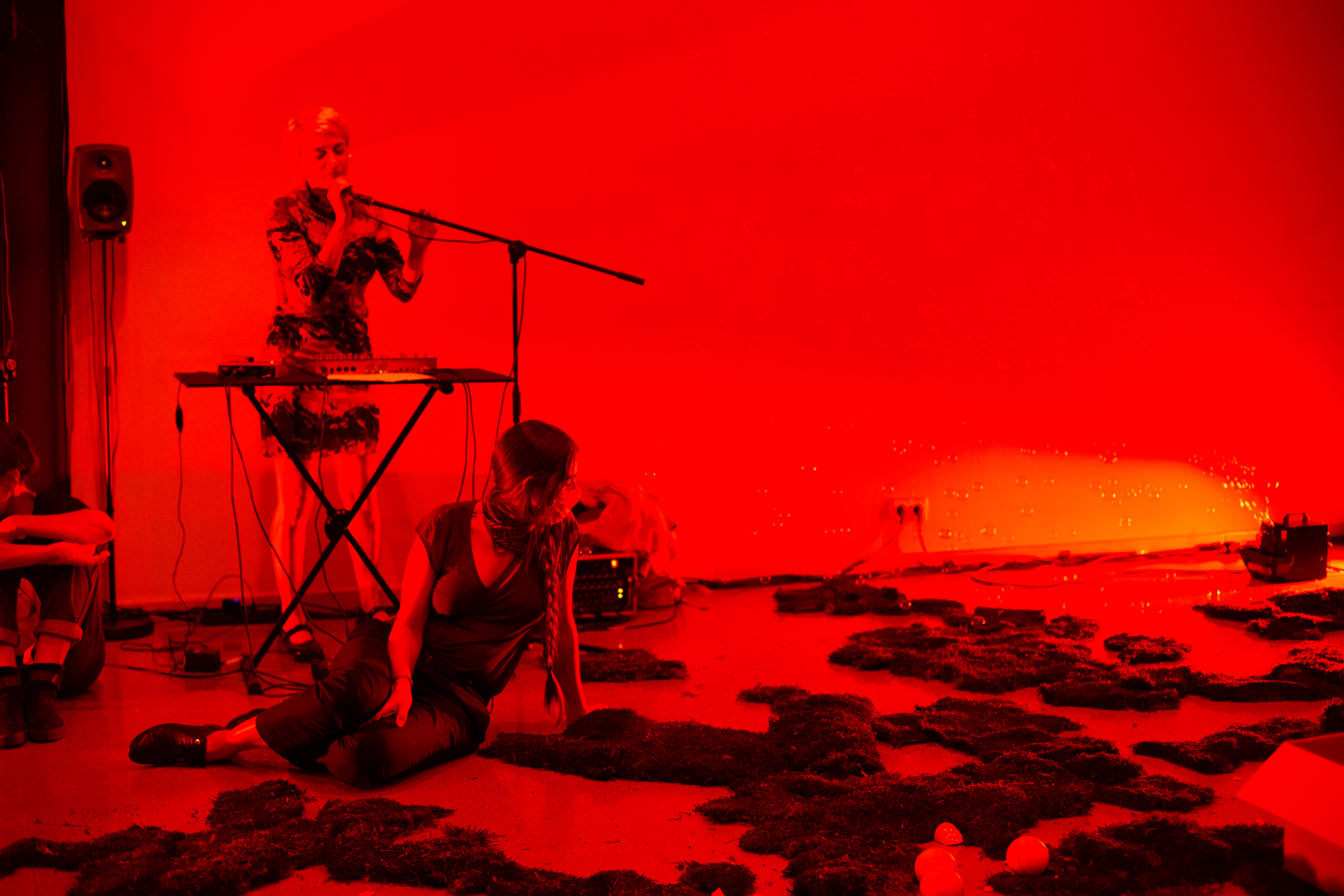 Ein in rotes Licht getauchter Raum. Auf dem Boden sitzt eine Person zwischen ringsum verteilten Moosen. Im Hintergrund ist eine weitere Frau an einem Keyboard zu sehen.