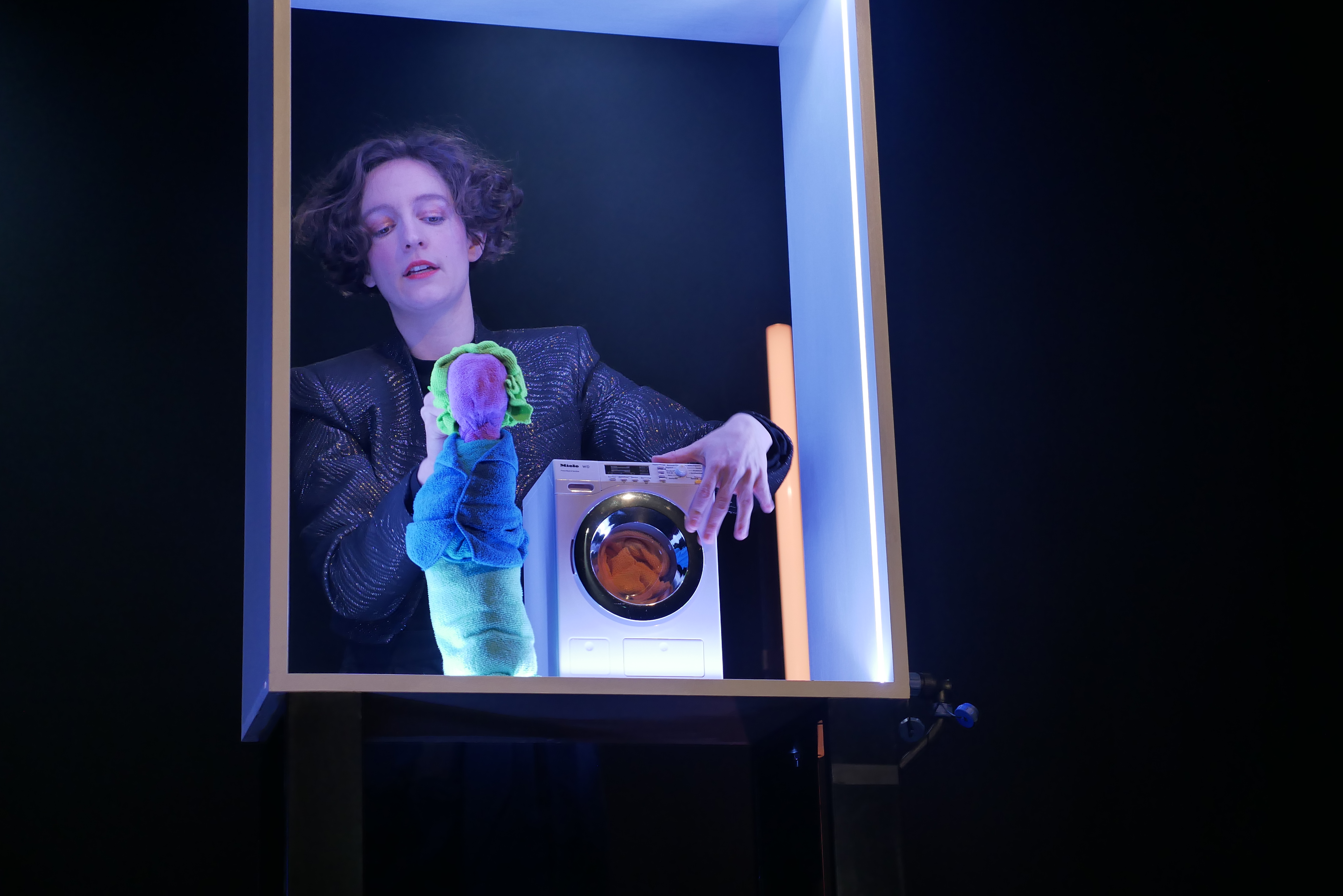 Josephine Hock hält die gesichtslose Stoffminiatur einer Person mit Rock, Pullover und grünen Haaren in der Hand. Die Puppe steht neben einer Miniaturwaschmaschine.