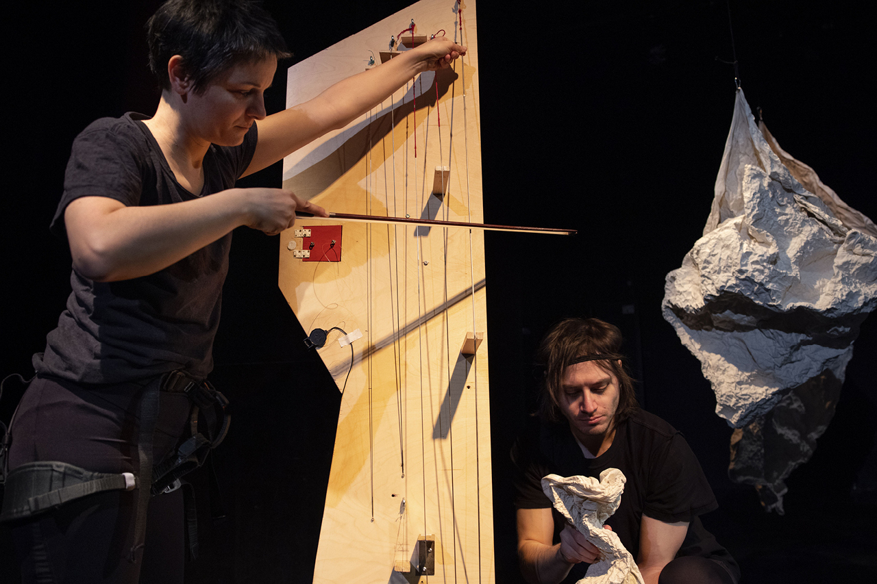 Performerin steht und streicht mit Geigenbogen die Saiten eines selbstgebauten Holzinstruments. Rechts daneben hockt ein weiterer Spieler, der mit den Händen ein papierartiges Material animiert als ob es eine Puppe wäre.