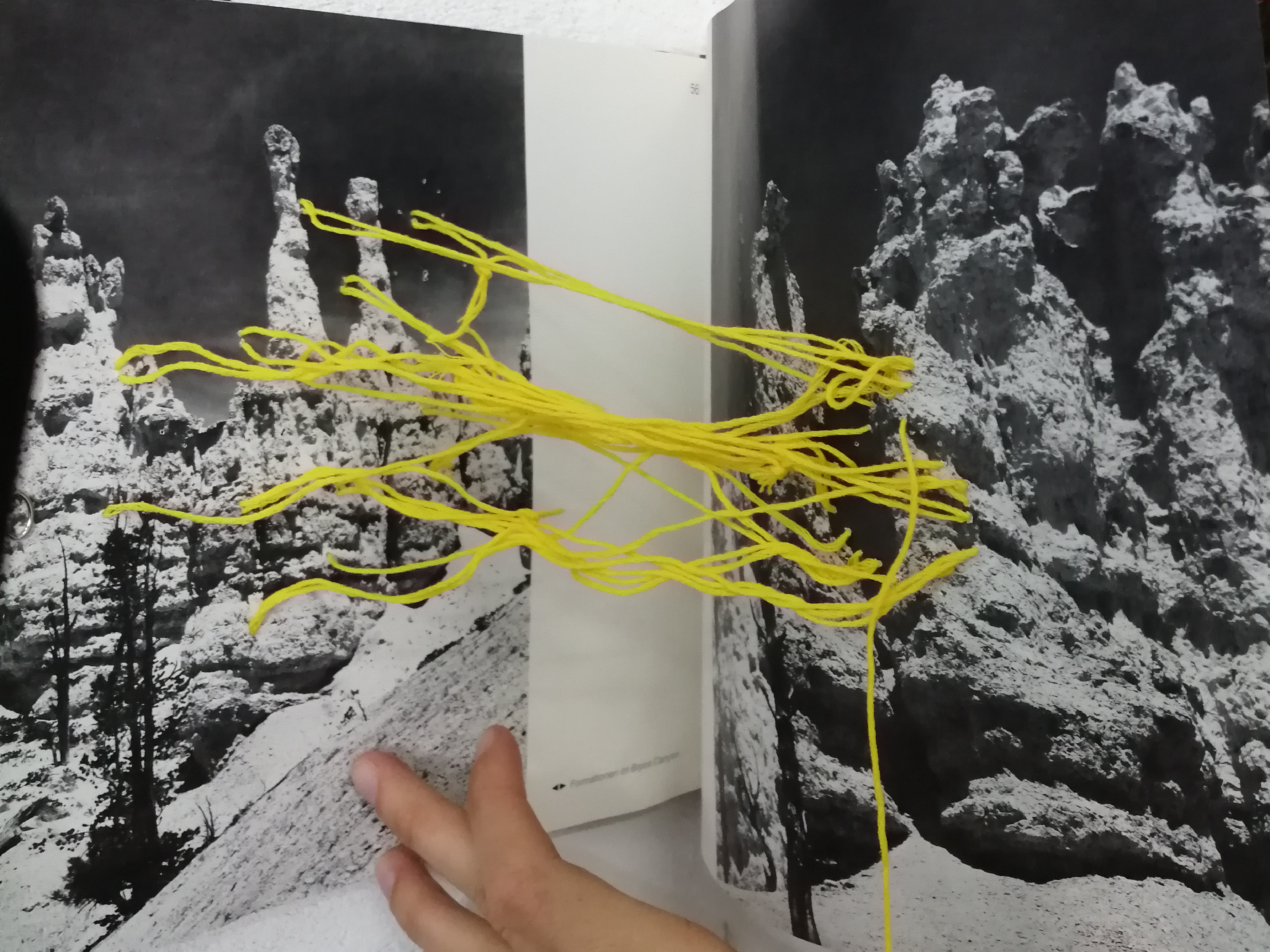 Die sich gegenüberliegenden Seiten eines Buchs sind mit gelber Schnur an verschiedenen Punkten fixiert und miteinander verbunden. Die Seiten sind mit schwarz-weiß Fotos von Gesteinsformationen bedruckt.