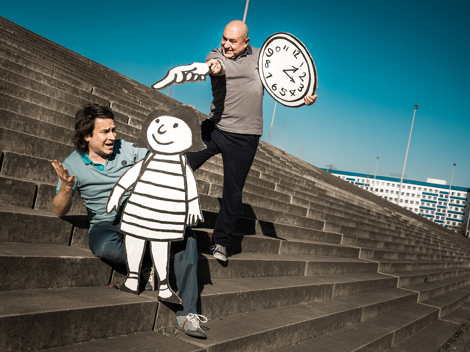 Zwei Spieler mit einer lebensgroßen Flachfigur und einer Uhr aus Papier auf Treppen posierend.