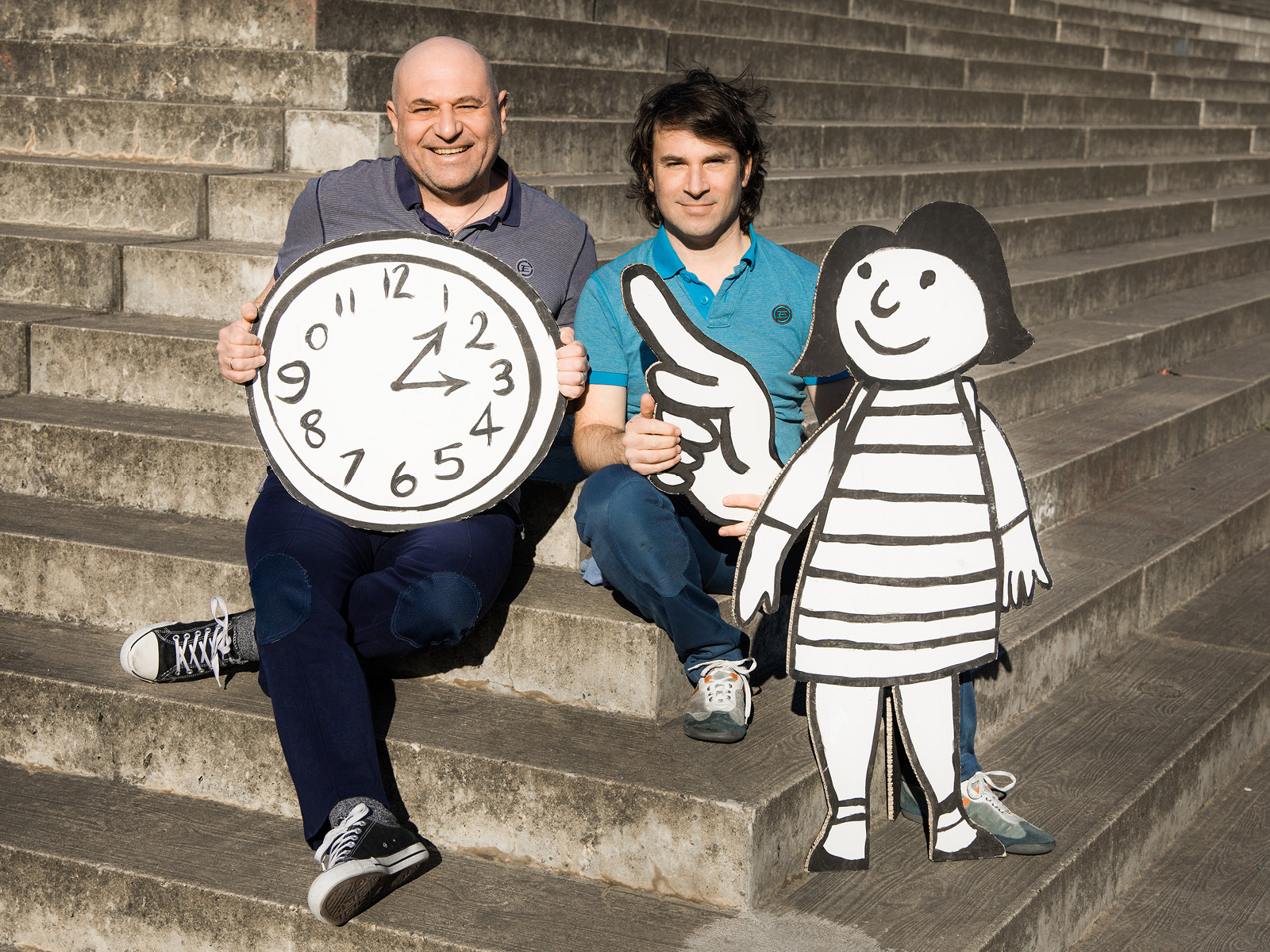 Zwei Spieler mit einer lebensgroßen Flachfigur und einer Uhr aus Papier auf Treppen sitzend.