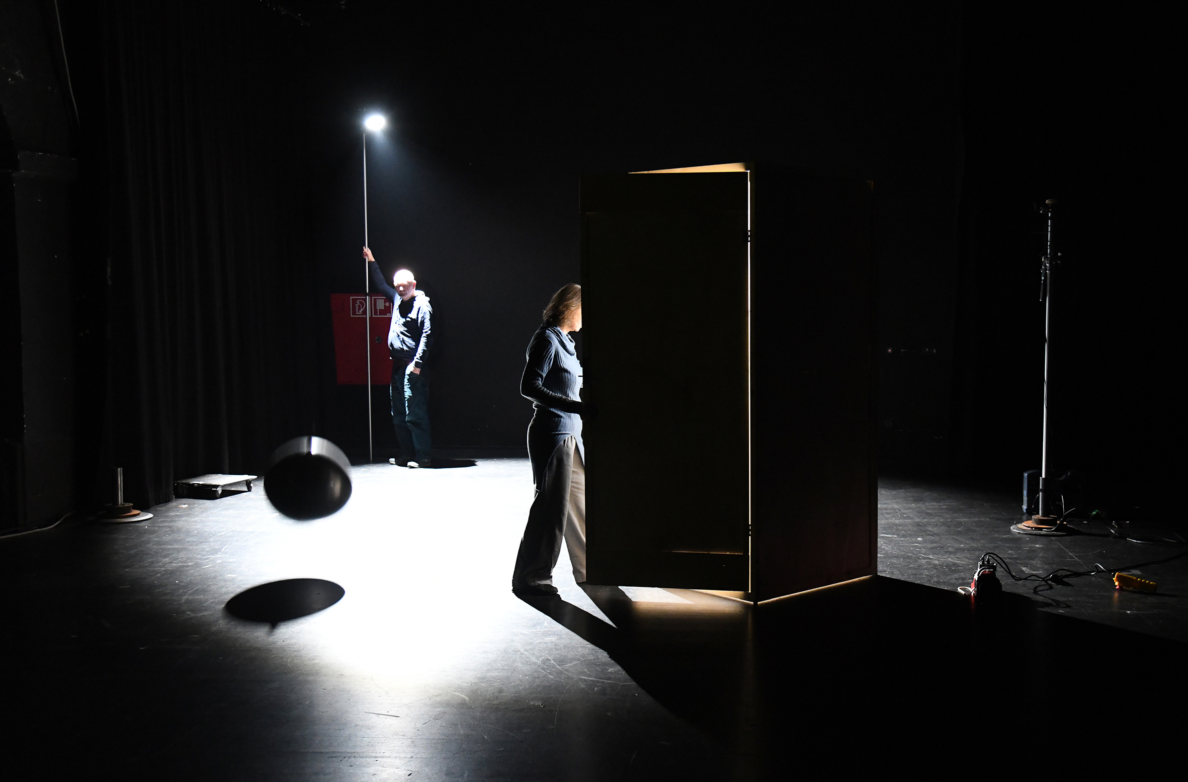 Zwei Personen im dunklen Bühnenraum, eine geht gerade in eine von innen beleuchtete Holzkiste. Die hintere Person hält eine Lampe, deren runder Lichtkegel auf dem Bühnenboden strahlt.