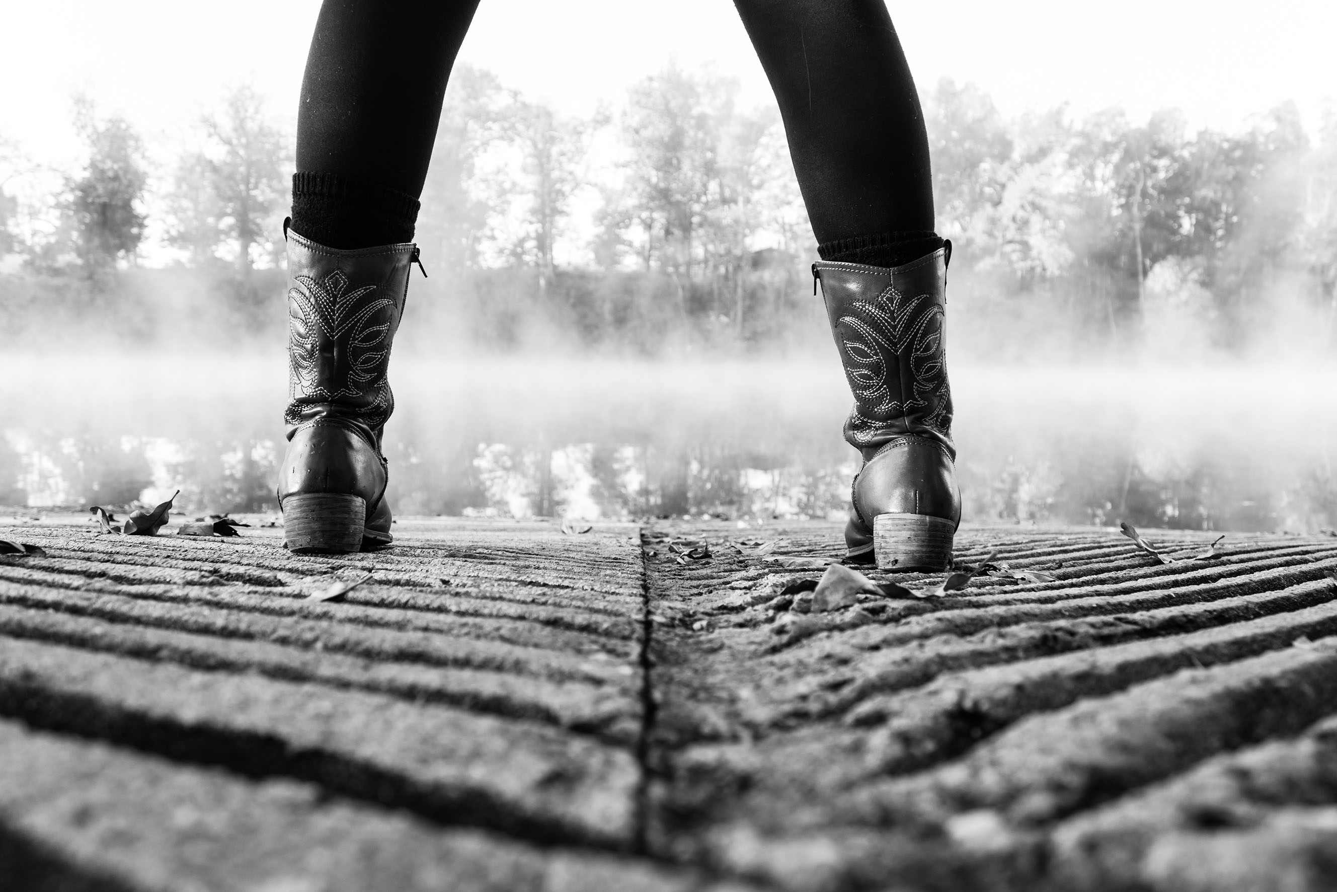 The black and white photo shows the lower legs of a woman in black pants. She wears cowboy boots and stands on an uneven earth surface.