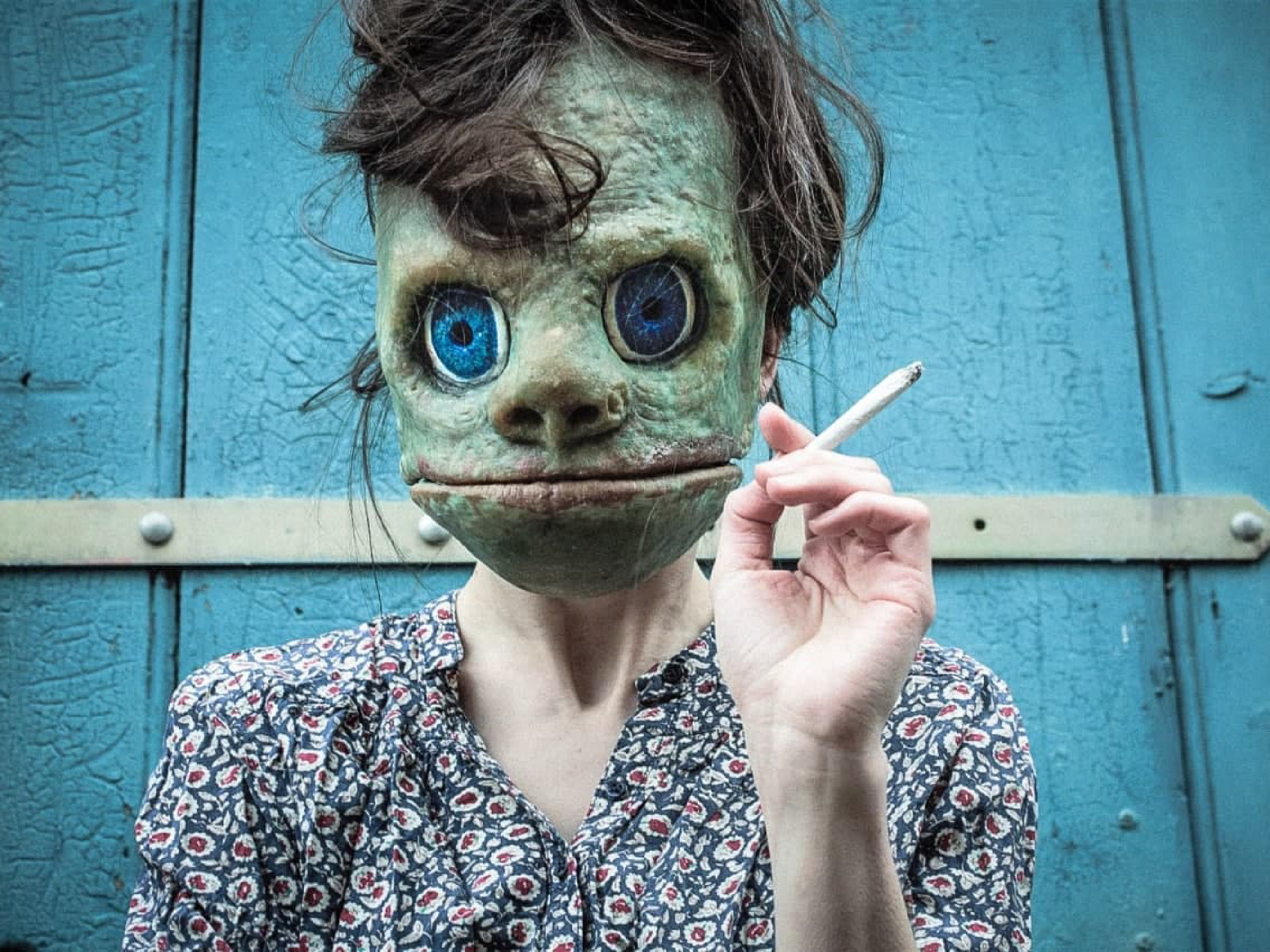 Eine Frau in Blümchenkleid trägt eine grüne Maske mit breitem Mund und großen, blauen Augen. Sie hält eine Zigarette in der Hand. Im Hintergrund ist eine Wand aus hellblauen Latten zu erkennen.