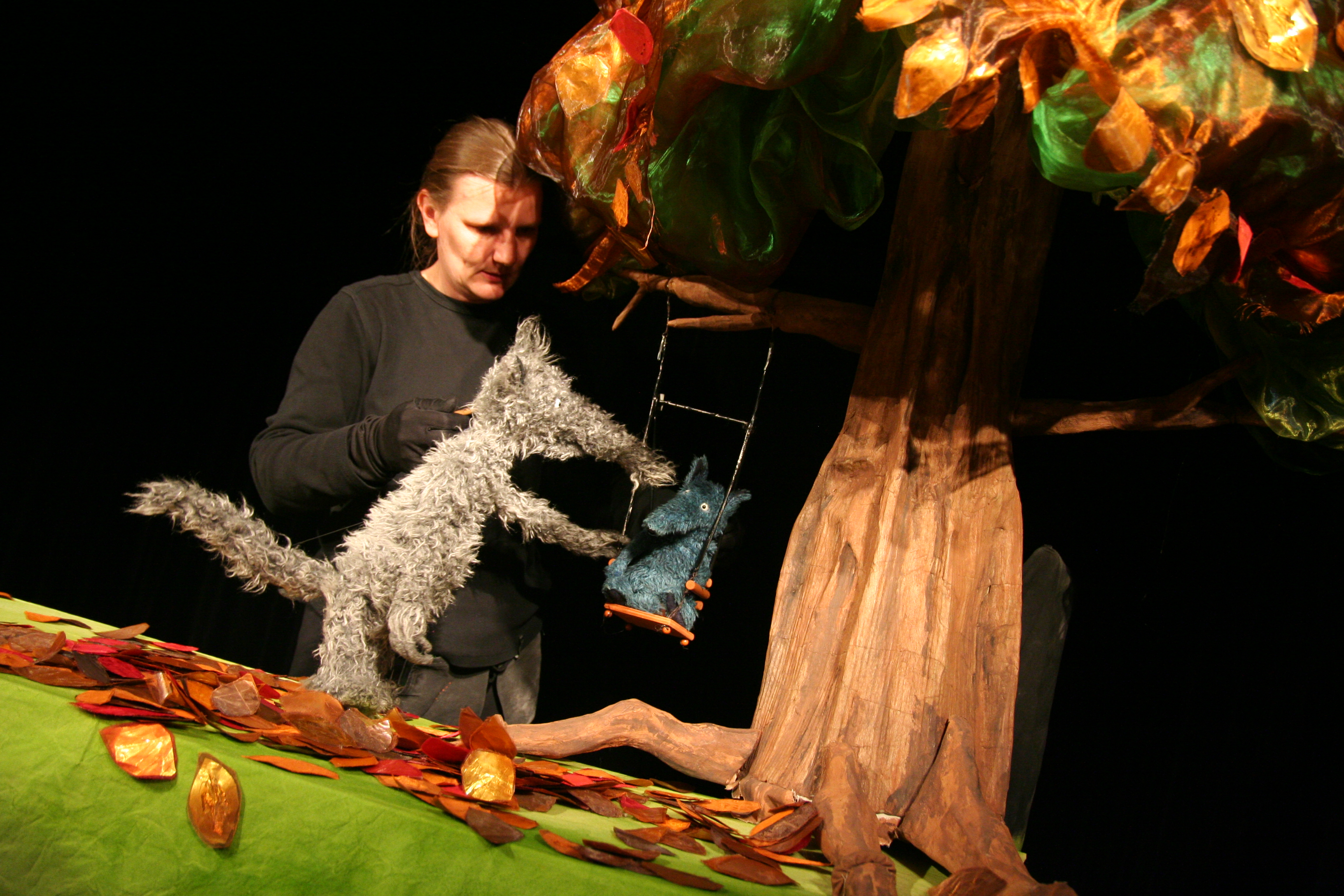 Die Figur eines grauen Wolfs steht unter einem großen Baum mit Herbstlaub. An dem Baum ist eine Schaukel befestigt, auf der ein kleiner, blauer Wolf sitzt. Die beiden Wölfe werden von einer dunkel gekleideten Puppenspielerin im Hinergrund geführt.