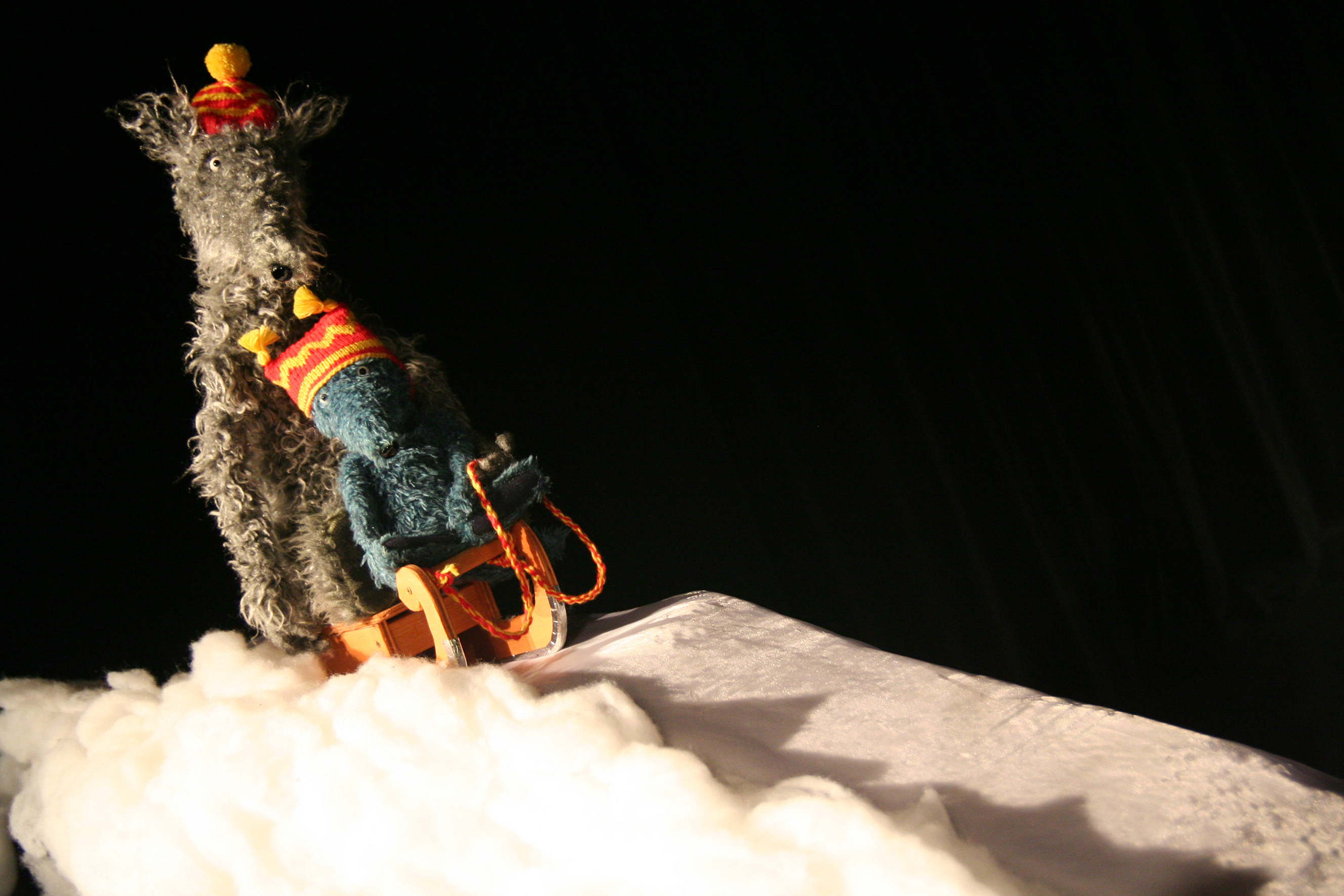 Two figures, a grey wolf and a smaller, blue wolf with colorful bobble hats stand in a winter landscape. The smaller wolf is sitting on a sledge.