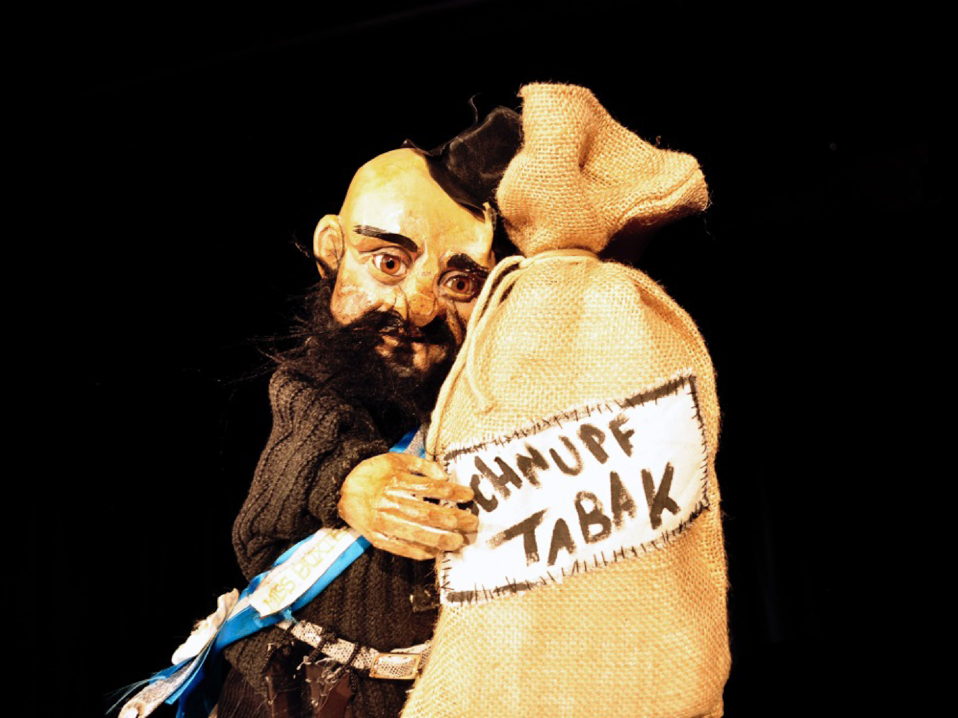 The puppet of the robber Hotzenplotz wears a black top hat on his bald head. He is hugging a bag labeled Snuff.