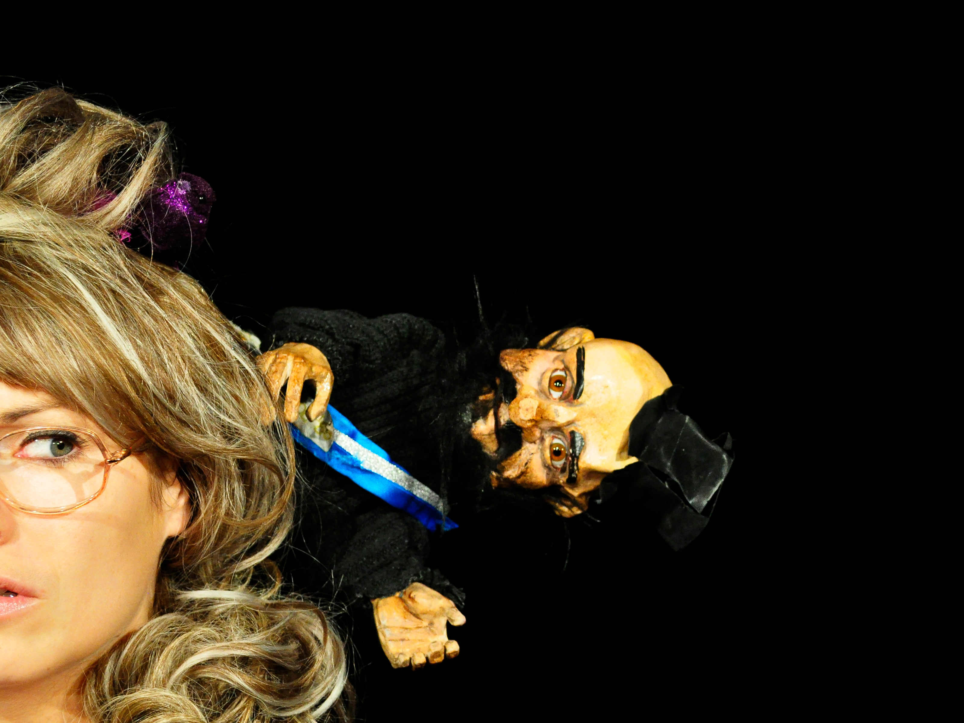The face of a middle-aged woman with a blonde wig and rimless glasses is half-cut. On her head sits the doll of a darkly dressed man with a top hat and beard.
