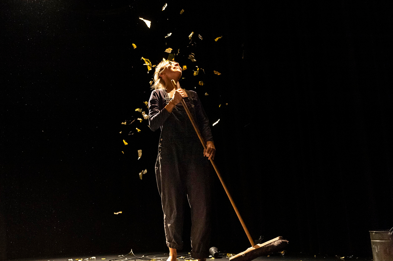 Performerin mit Besen in  der Hand auf dunkler Bühne. Über sie regnet es goldene Folienflocken. Am rechten Bildrand sieht man ein Stück von einem Eimer.