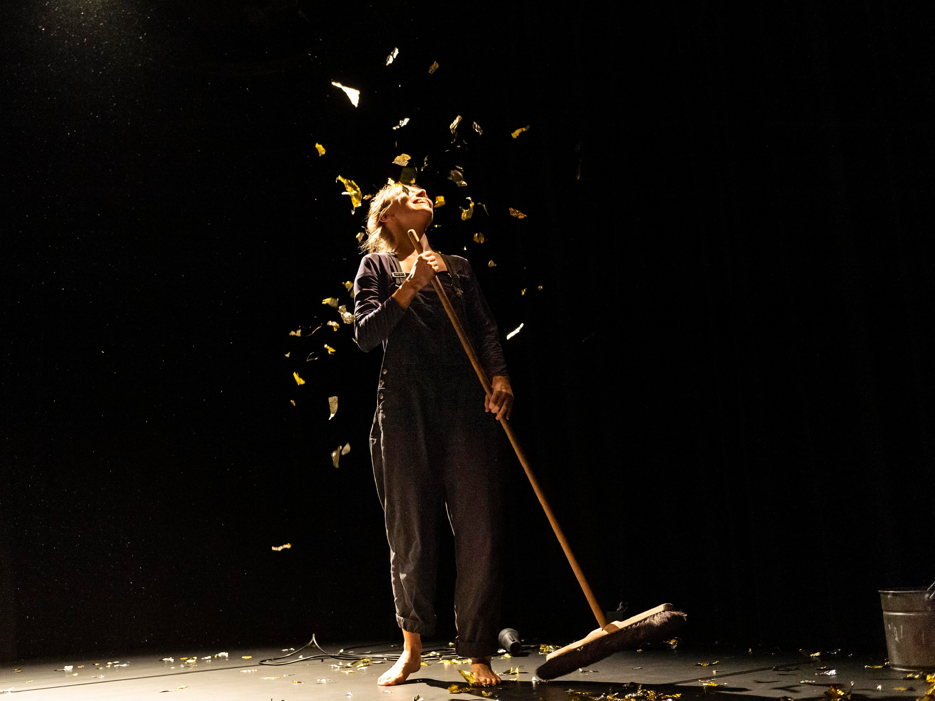 Gold confetti rains down from the ceiling onto the darkly lit stage on player Karoline Hoffmann. She raises her head to the confetti and holds a long-handled broom in her hand.