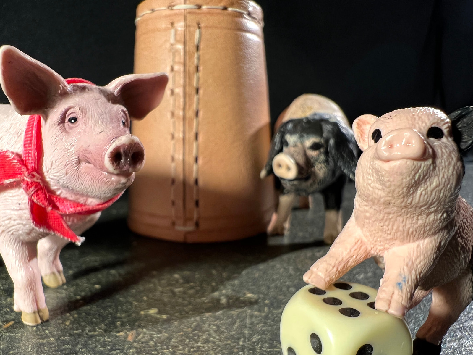 Die Figur eines kleinen Schweinchens stützt die Pfoten auf einen Würfel mit der Punktzahl sechs. Im Hintergrund sind noch zwei weitere Schweinfiguren, eines davon ist rosa und trägt ein rotes Halstuch. Das andere hat Schlappohren und einen schwarzen Kopf und befindet sich neben einem Würfelbecher aus Leder.