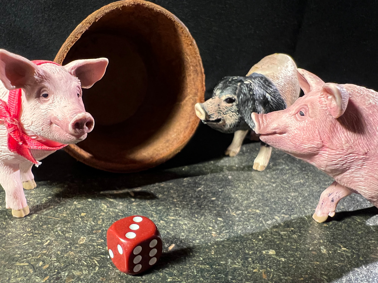 Die Figuren von drei Schweinchen, zwei rosa, eins braun mit Schlappohren umringen einen Würfel. Im Hintergrund liegt ein Lederbecher.