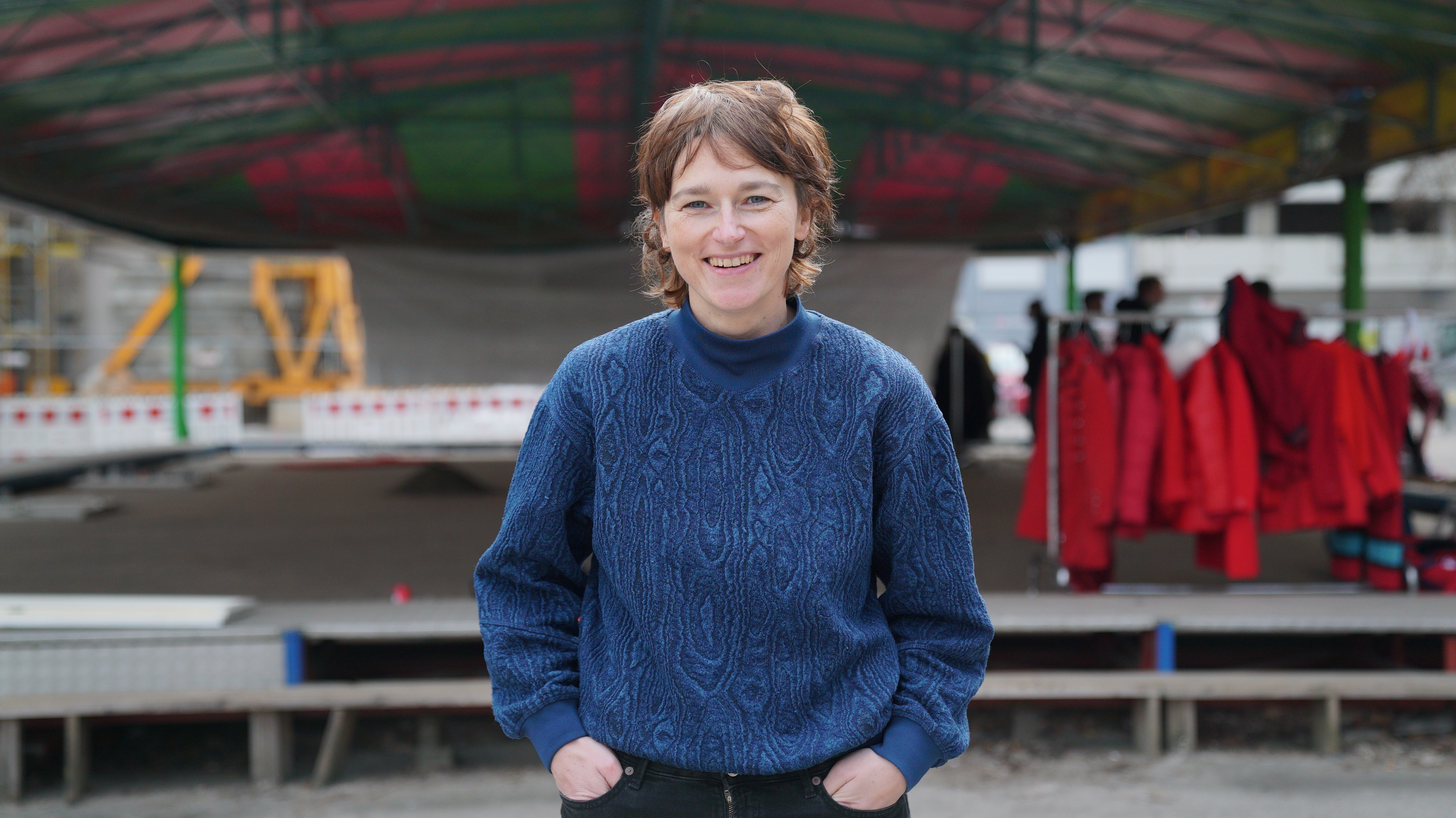Melanie Hinz has about shoulder-length brown hair and bangs and stands in front of an empty stage in a blue knitted sweater. She has her hands casually in her pockets and smiles broadly at the camera.