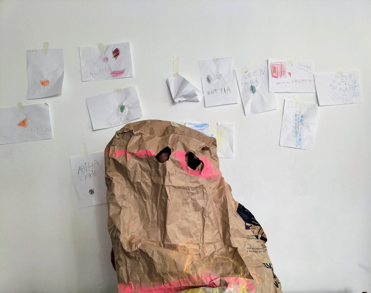 Melanie Hinz wears a brown, crumpled cardboard bag over her face. It looks like a stone and only has small holes cut into it for the eyes and nose. Behind her hang pictures of children.