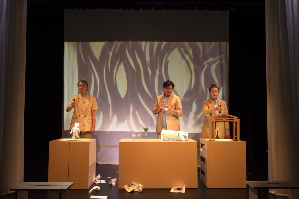 Nebeneinander stehen 3 Schauspieler vor je einem abgetrennten Teil eines Tisches, auf dem Figuren bzw. das Modell eines Theaterhauses stehen. Im Hintergrund Projektionen.