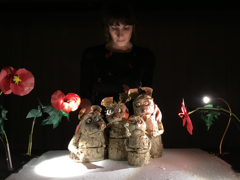 Vier Mäusefiguren sitzen auf einer Styroporplatte. Sie sind umgeben von großen Papiermohnblumen. Gehalten werden sie von Puppenspielerin Sabine Mittelhammer. Ihr Gesicht ist leicht von unten beleuchtet. Der Hintergrund ist schwarz und dunkel.
