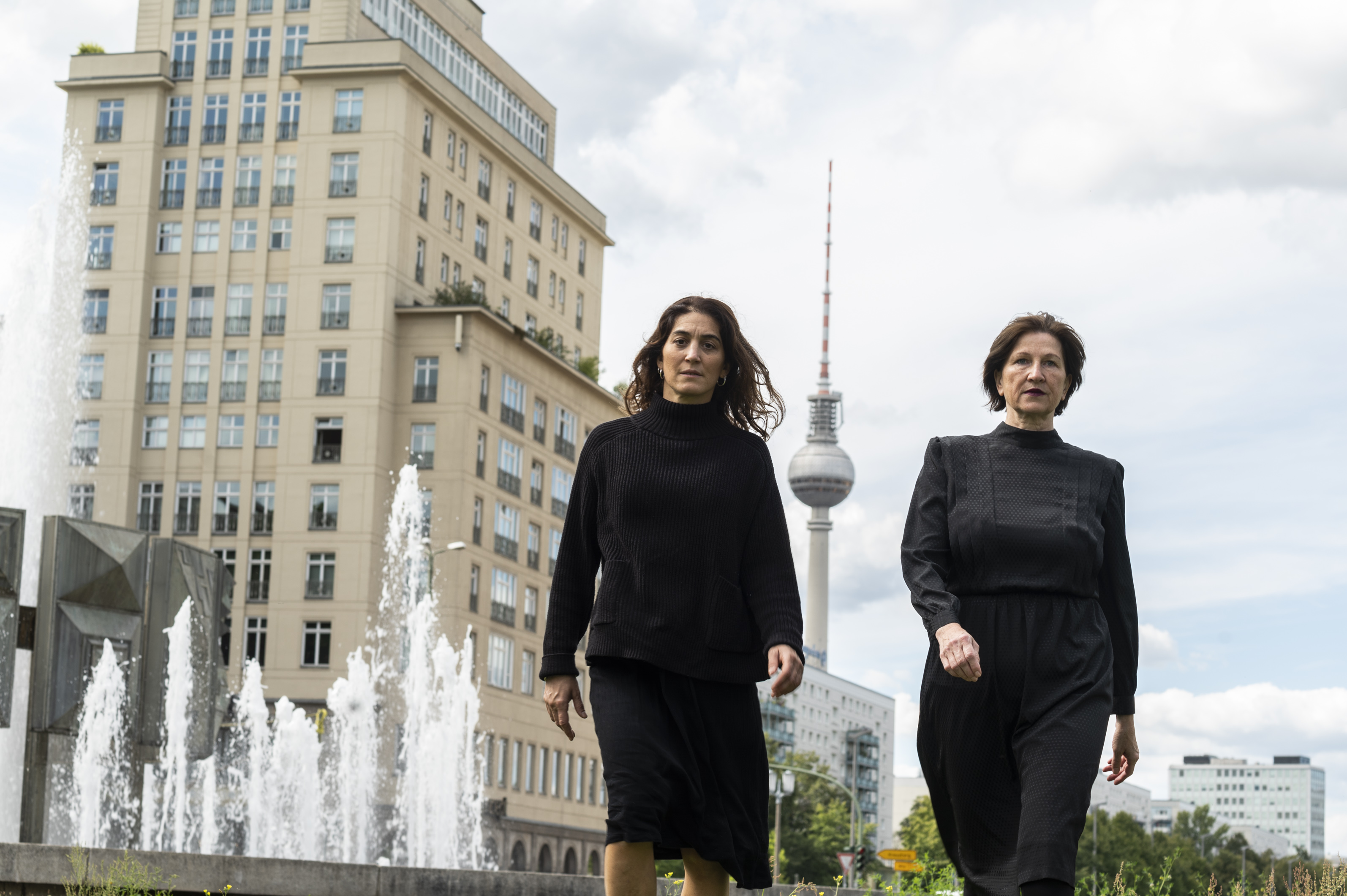 Zwei schwarz gekleidete Frauen, diesmal unmaskiert, laufen durch Berlin. Hinter ihnen ist der Fernsehturm zu sehen.