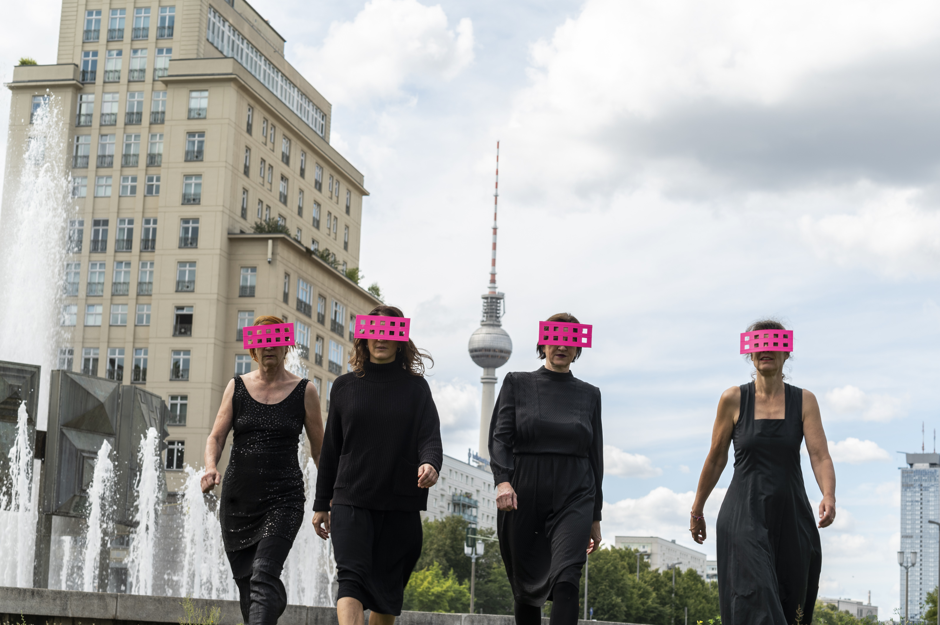 Vier Frauen laufen schwarz gekleidet durch Berlin. Hinter ihnen ist der Fernsehturm zu sehen. Sie tragen pinke Balken vor den Augen, die durchlöchert sind.