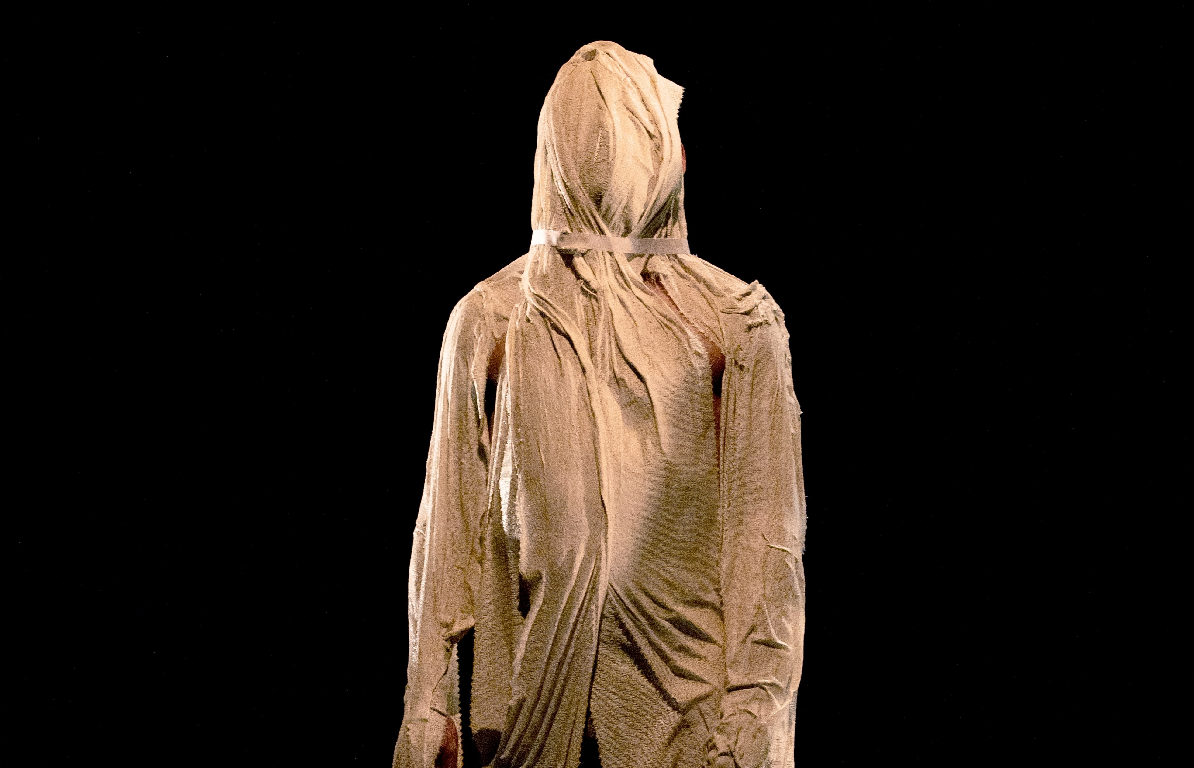 Der Körper und Kopf einer stehenden Person ist komplett mit einem eng anliegenden, falten werfenden Stoff überzogen. Er sieht dadurch aus wie eine Marmor Statue.
