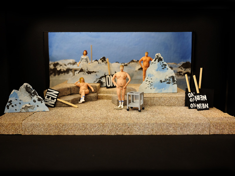 Drei nackte, menschliche Knetfiguren stehen in einer Landschaft aus Papierbergen. Im Hintergrund ist eine weibliche Knetfigur mit Kleid. Im Bild liegen vier Schilder. Auf dem vordersten Schild steht NEIN NEIN.