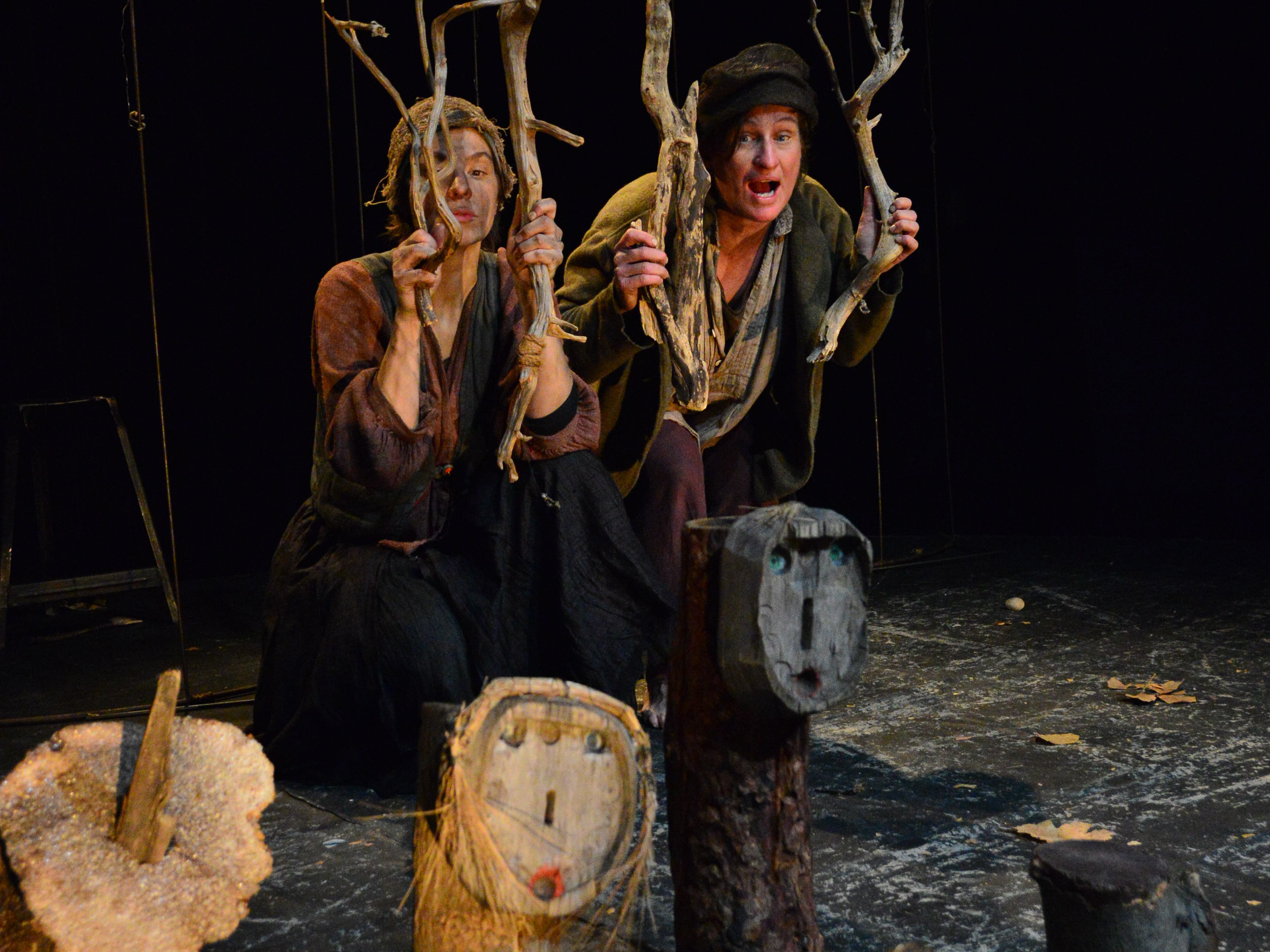 Hansel and Gretel stand with dirty faces behind a few wooden branches that suggest a cage. In front of them are two pieces of tree trunk with minimalist drawn faces.