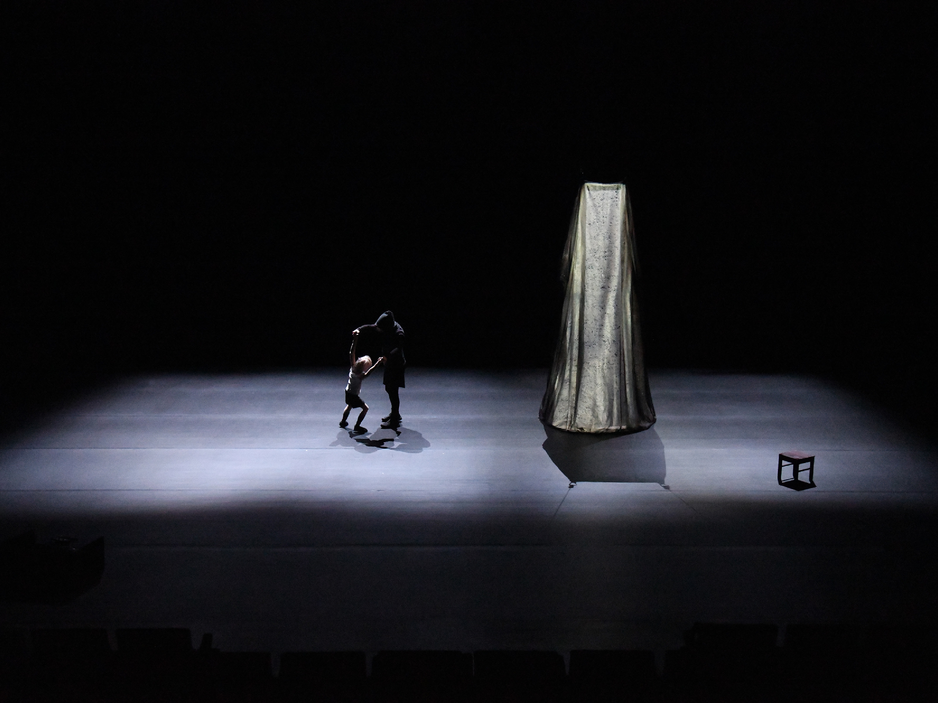On a diffusely lit stage, a human, all in black with a hood over his head, dances with a small boyish doll. The stage also includes a cylindrical falling shimmering fabric and a small stool.