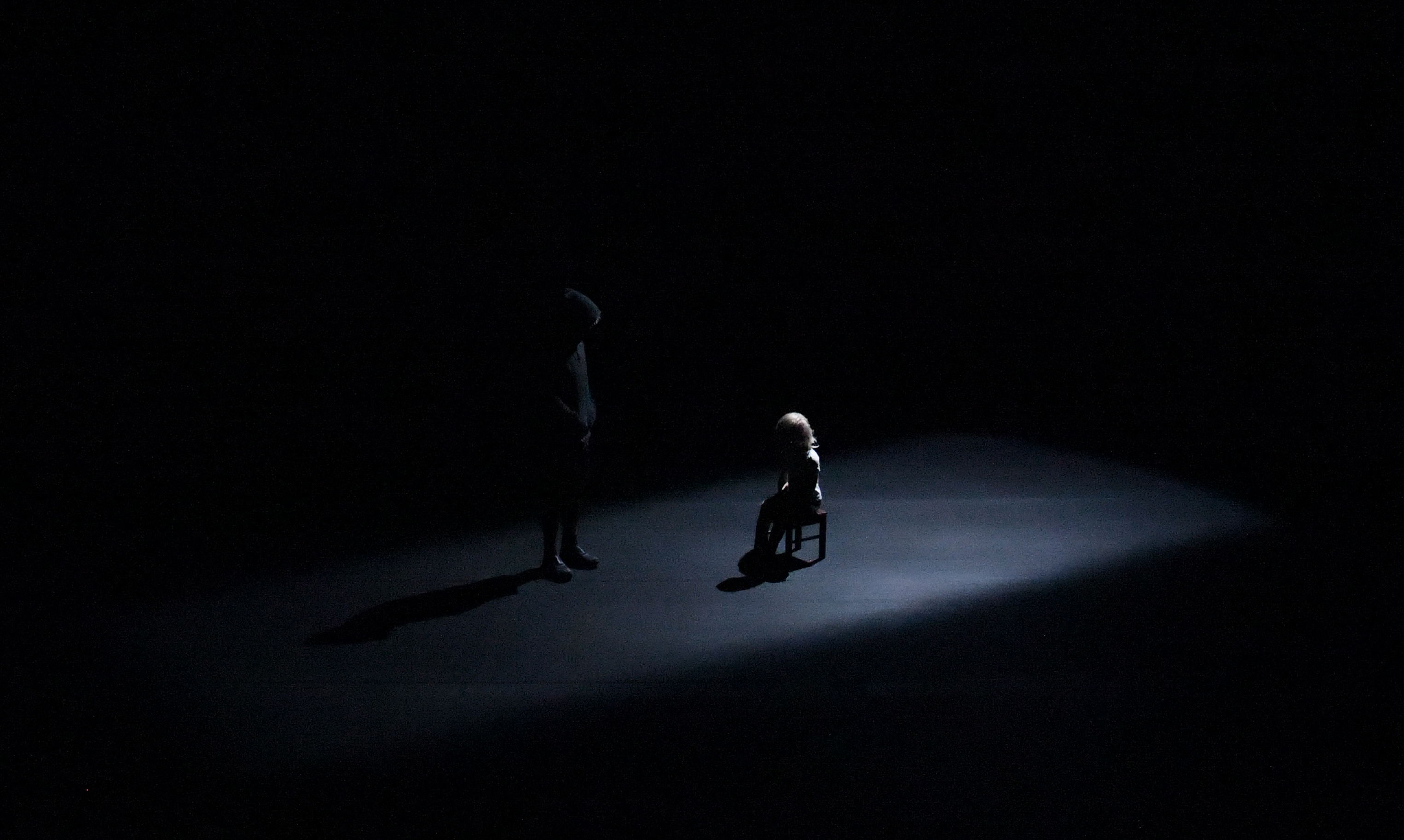 Puppe sitzt auf einem Höckerchen im spärlich beleuchteten schwarzen Bühnenraum. Links daneben steht der Spieler, durch die schwarze Kleidung fast unsichtbar in dem Dunkel.