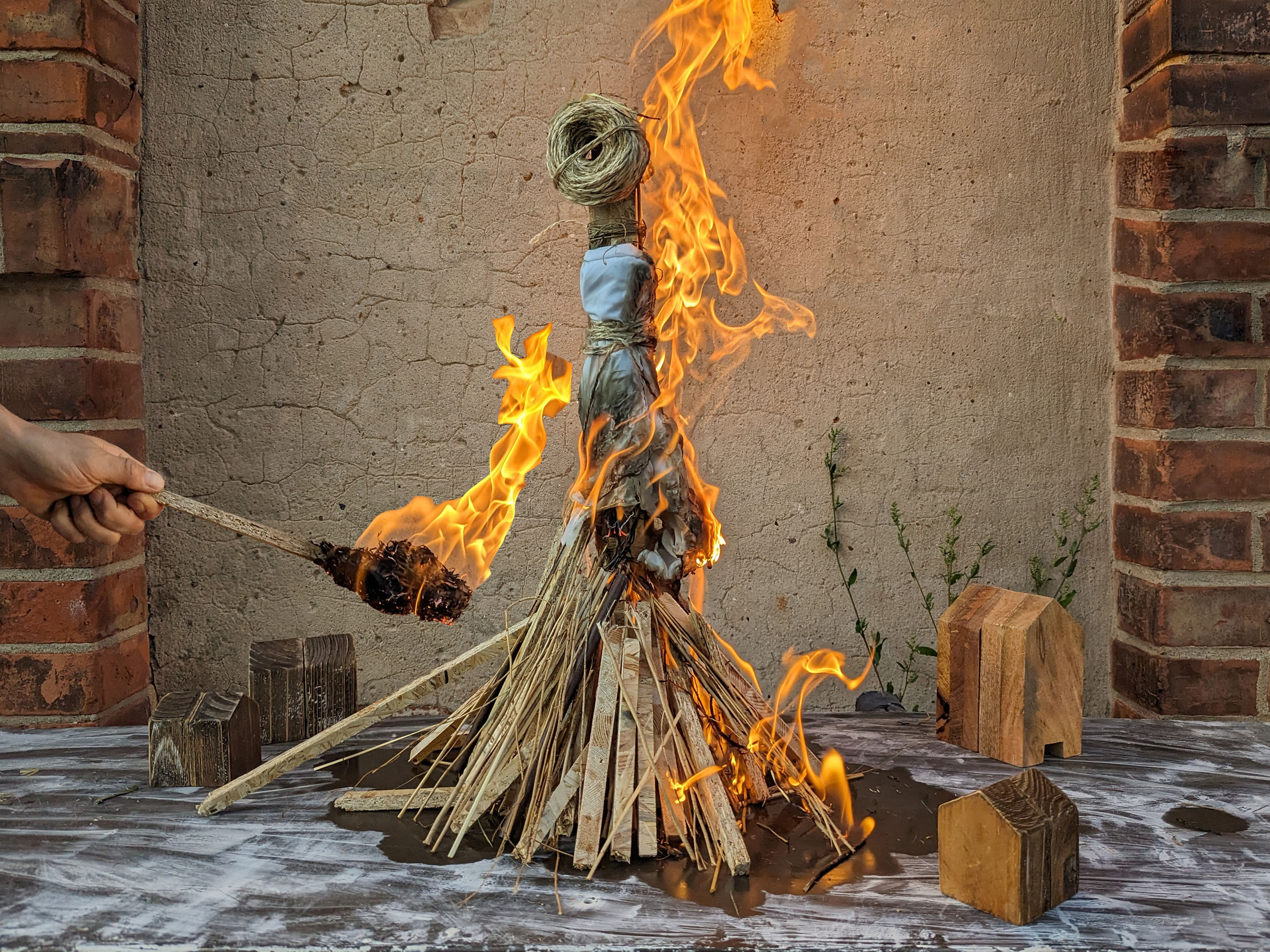 Von links ragt eine menschliche Hand mit Holzfackel ins Bild. Sie zündet einen Holzhaufen mit einer Strohpuppe an. Der Haufen und die Figur brennen lichterloh.