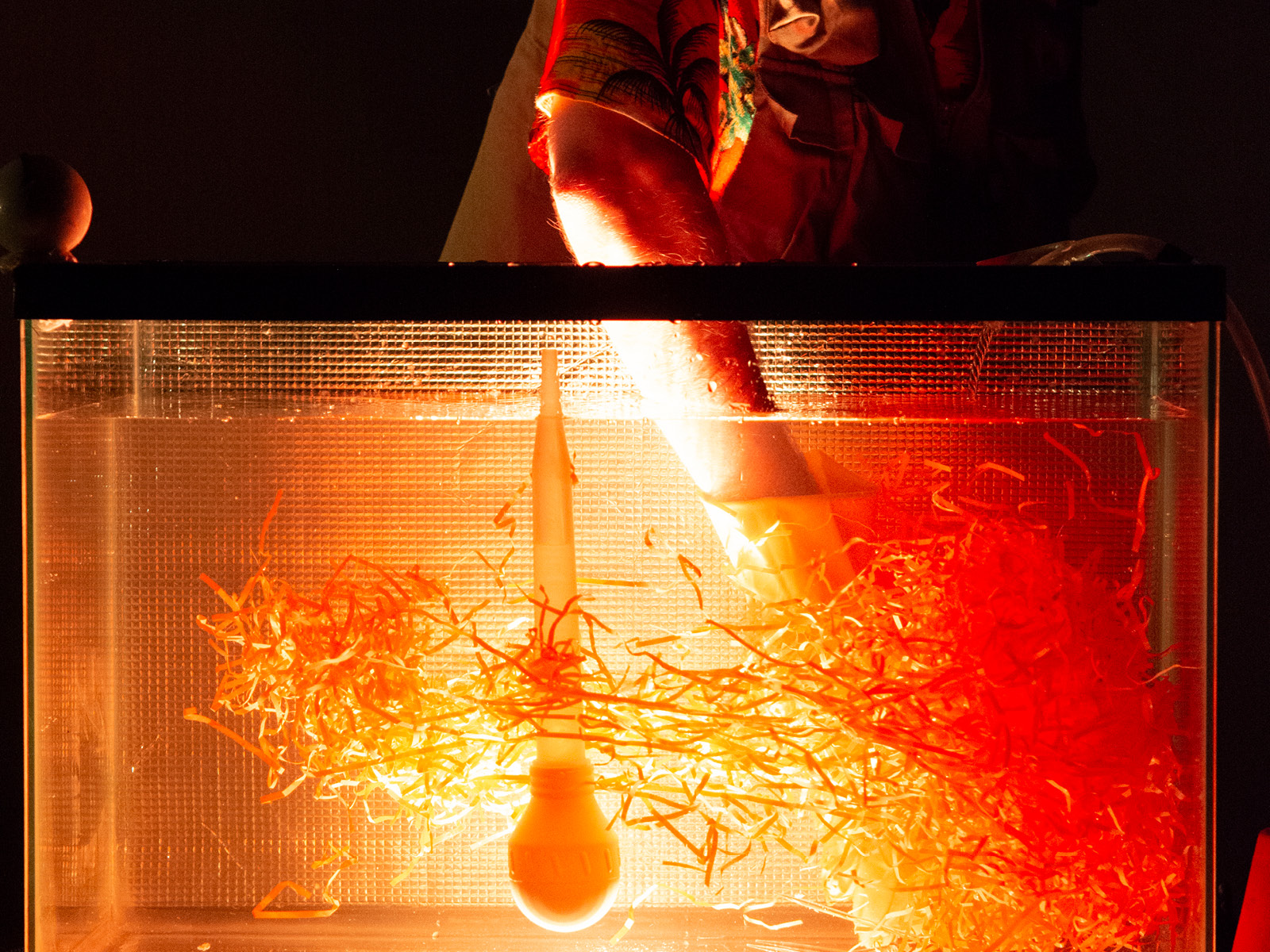Spaghettiähnliche in alle möglichen Himmelsrichtungen ragende Strohhalme und eine große Pipette mit Gummipfropfen treiben in einer orange-rot beleuchteten Aquarium, in dem das Hand von Robin Leveroos hineinragt.