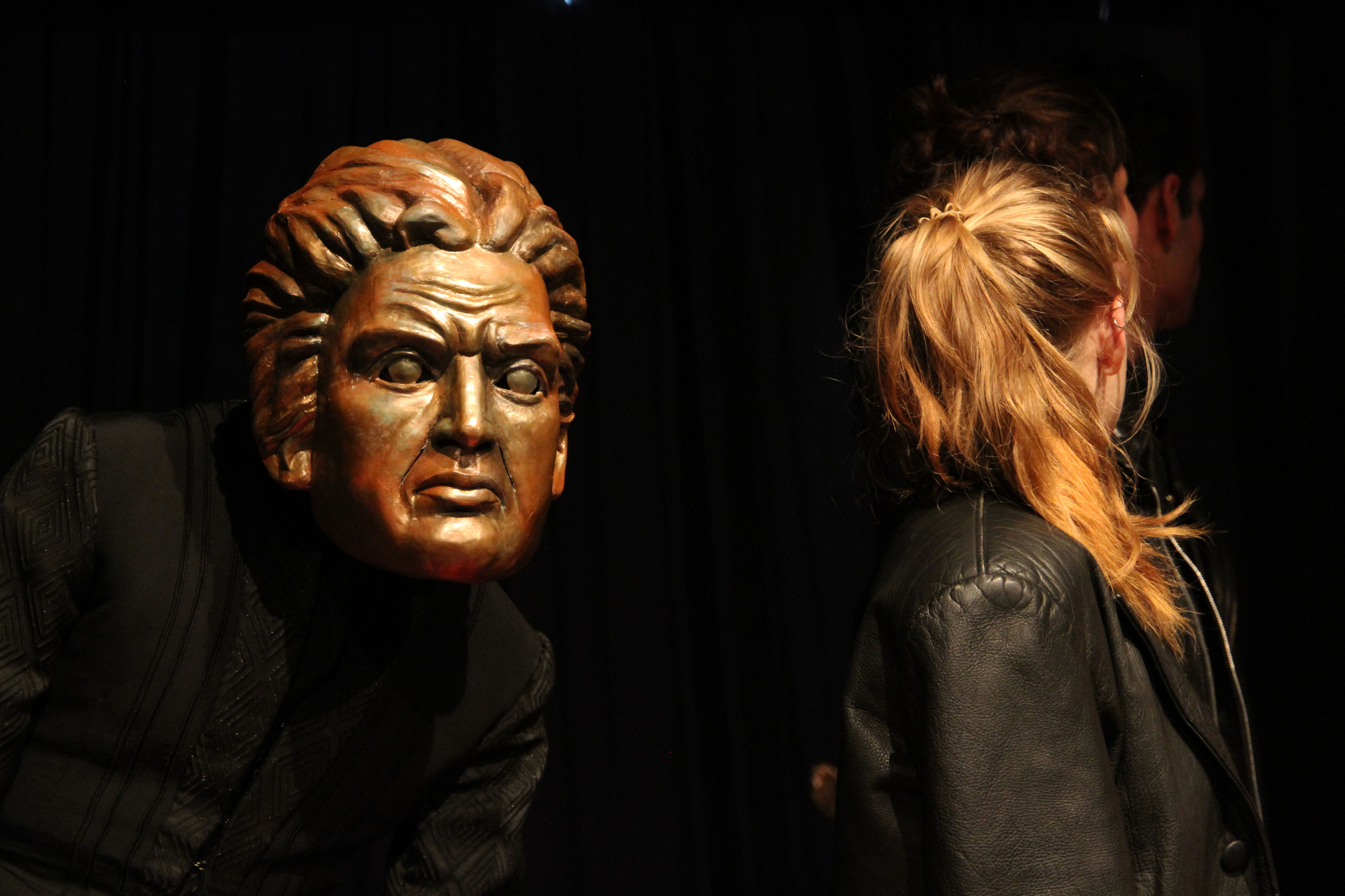 A man in black clothes wears a golden face mask with a furrowed brow. Next to him is a girl with blond hair. She has turned her head away from him.