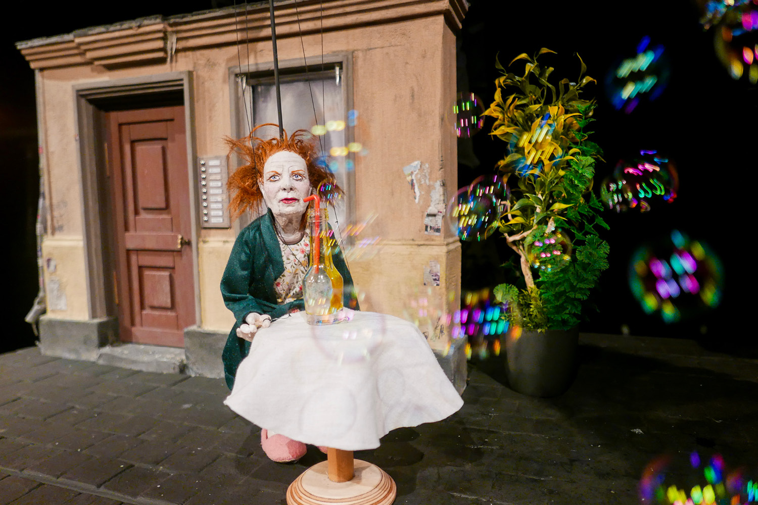 Eine rothaarige Marionettenfrau sitzt an einem runden Esstisch mit weißer Serviette und Kerze. Hinter ihr ist ein Haus mit einer Tür. Sie wird von Seifenblasen angepustet.