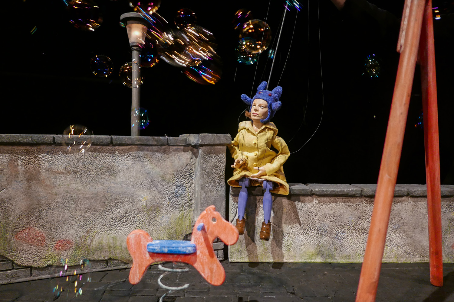 Eine Marionettenfrau mit Mütze und Regenmantel sitzt auf einem Spielplatzgelände mit Schaukelpferd und wird von Seifenblasen angepustet