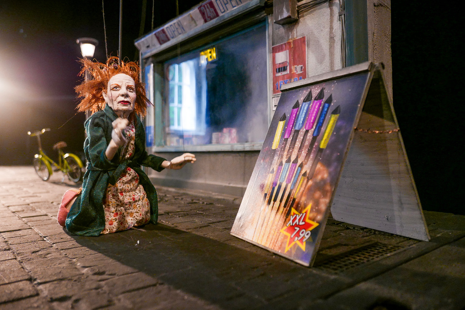 Eine rothaarige Marionettenfrau in Morgenmantel kniet neben einem Werbeschild mit Feuerwerksraketen. Im Hintergrund ist ein Fahrrand und ein Geschäft mit großem Schaufenster.