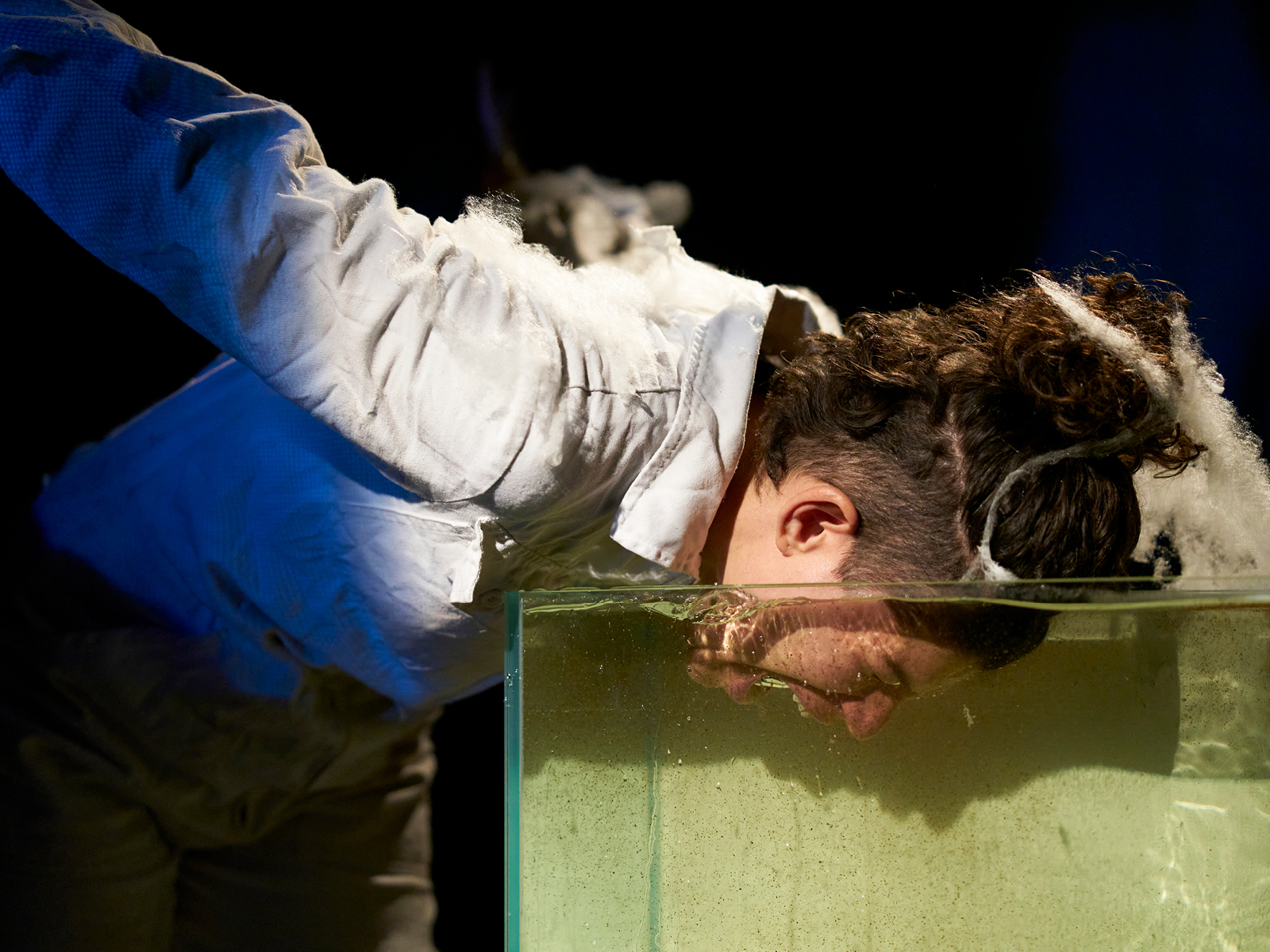 Performerin taucht ihren Kopf in ein mit Wasser gefülltes Aquarium. Ihr Mund ist weit geöffnet, als ob sie schreit.