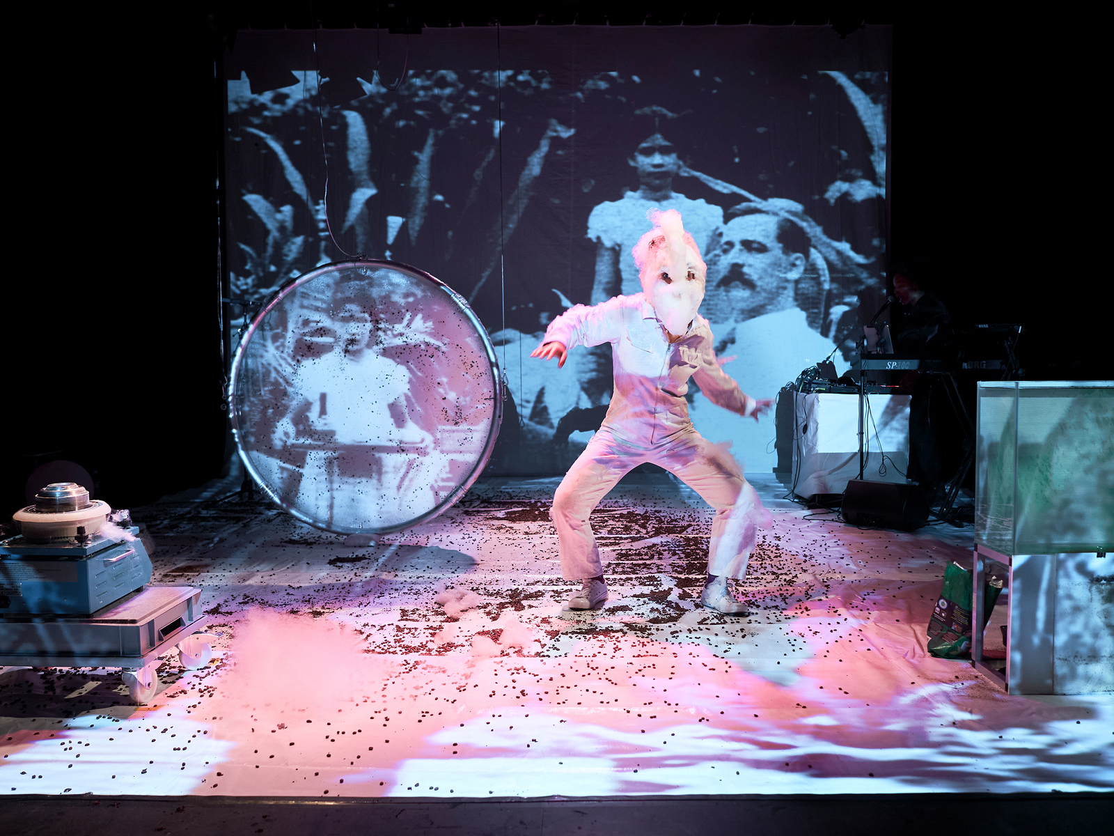 Auf der Bühne tanzt eine Performerin mit einer Maske aus Zuckerwatte vor einer Videoleinwand, auf die ein historisches Bild projeziert ist, das einen kolonial wirkenden Mann mit Schnurrbart zeigt, neben dem eine indigene Dienerin steht. Rechts im Bild ist ein Aquarium. Der Bühnenboden ist mit Kaffeebohnen bedeckt.