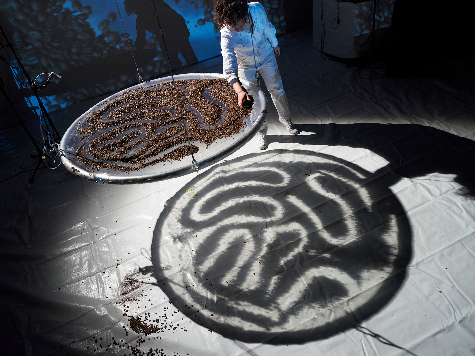 Auf einer an Drähten hängenden Schreibe sind Kaffeebohnen zu einem Muster zusammengeschoben. Darunter zeichnet sich das Schattenbild dieses Musters auf dem Boden ab.