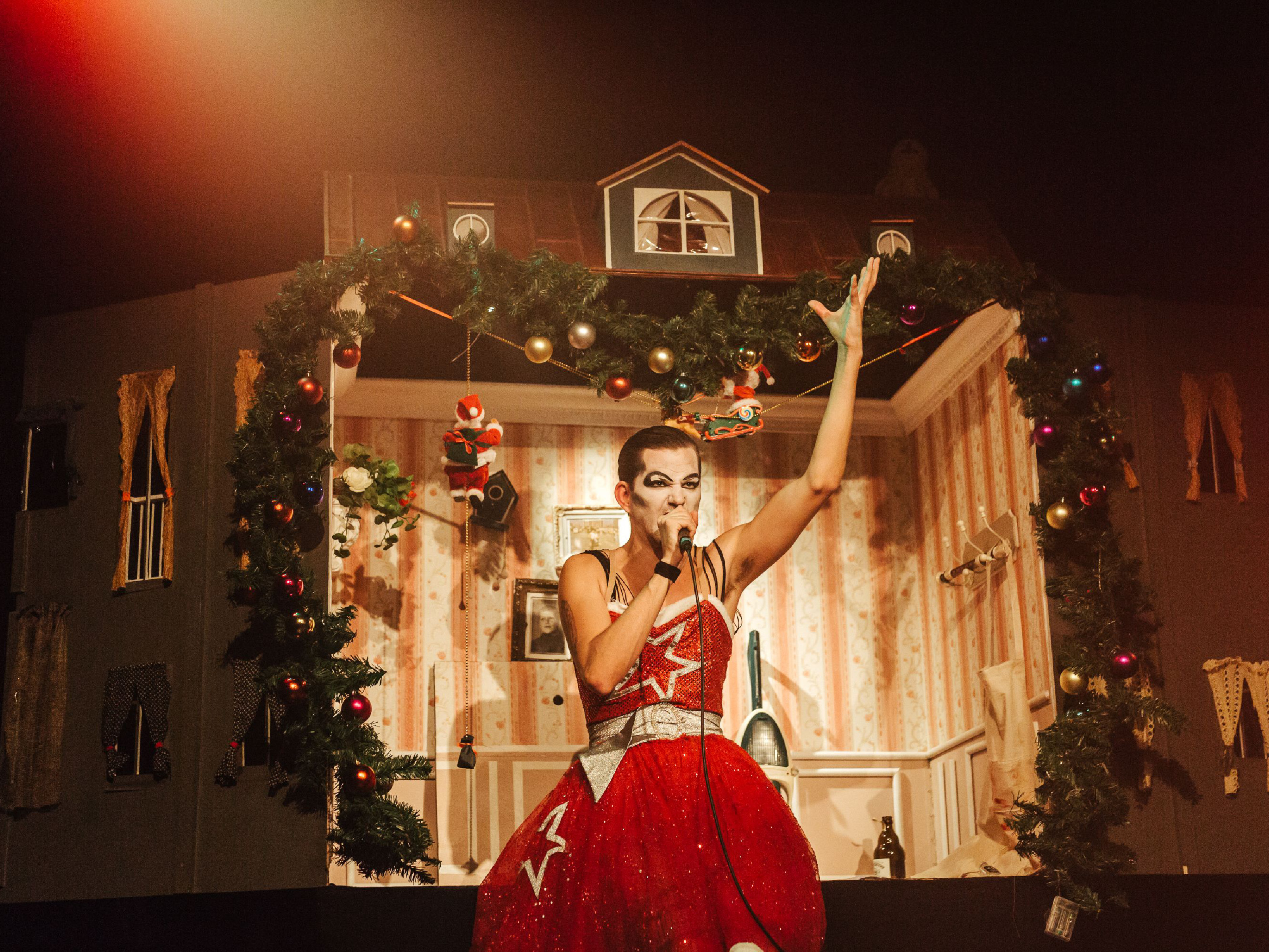 Performer Jarnoth stands in red party dress with glitter star and drag make-up in front of a large dollhouse room with Christmas decorations and sings passionately in a microphone he holds in his left hand. His right arm is stretched far upwards.