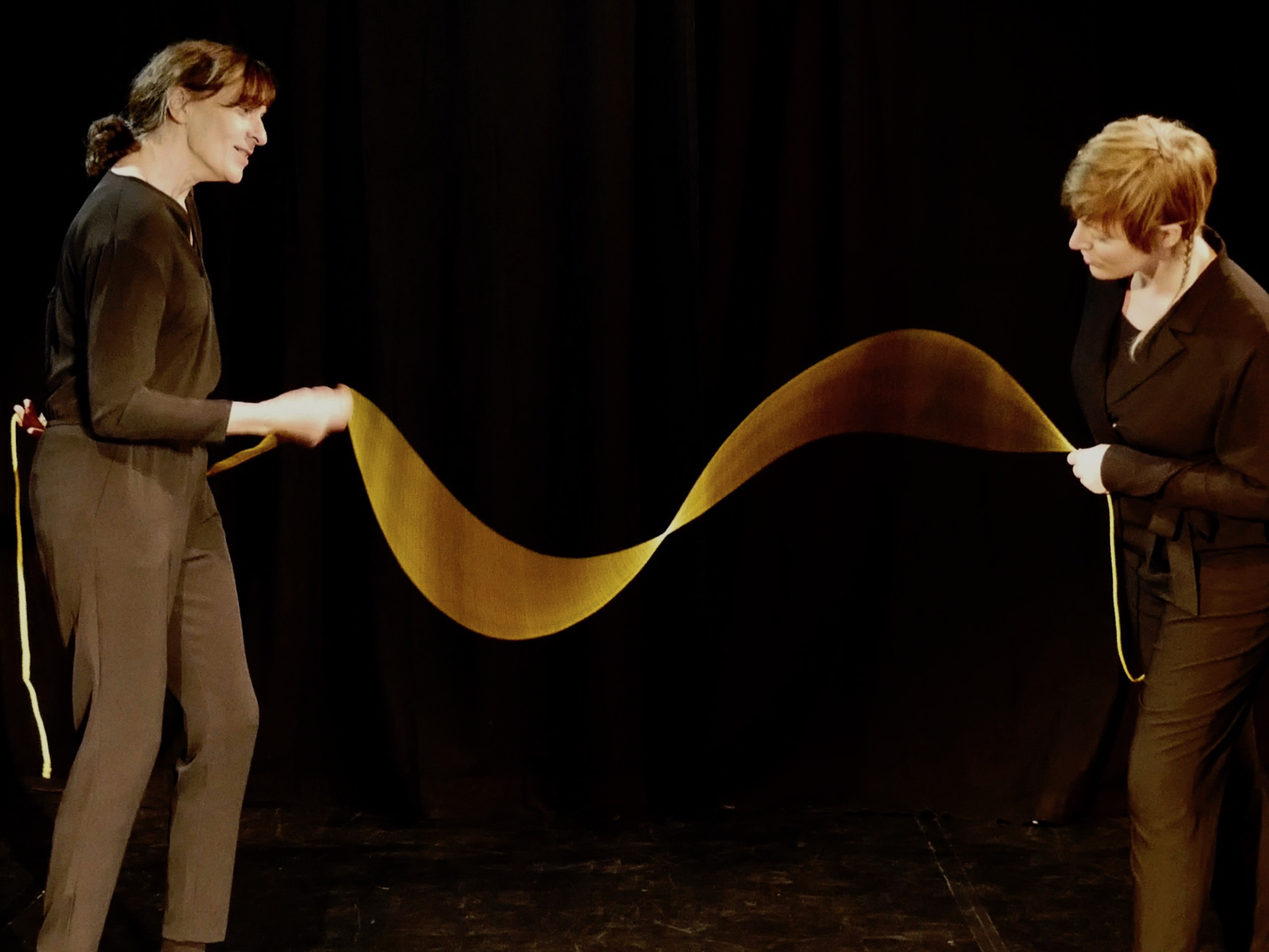 Zwei in Schwarz gekleidete Frauen stehen auf der Bühne und halten eine Art langes Band in der Hand, das zwischen ihnen wie eine Welle fließt.