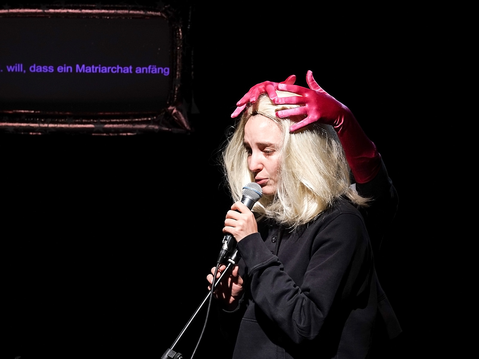 Eine blone Frau steht angespannt am Mikro. Aus dem Dunkel hinter ihr legen sich rosafarbene Samfthandschuhe auf ihren Kopf.