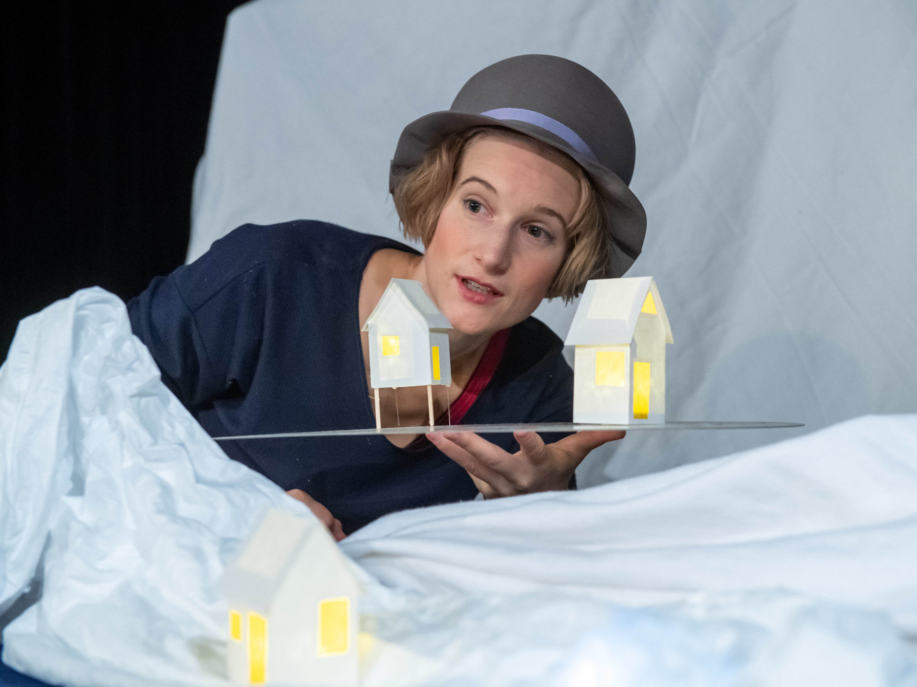 Figurenspielerin Miriam Hesse steht mit grauem Hut in einer verschneiten Landschaft und hält zwei kleine Häuschen auf einer Platte in der Hand. Aus den Häuschen scheint warmes Licht.