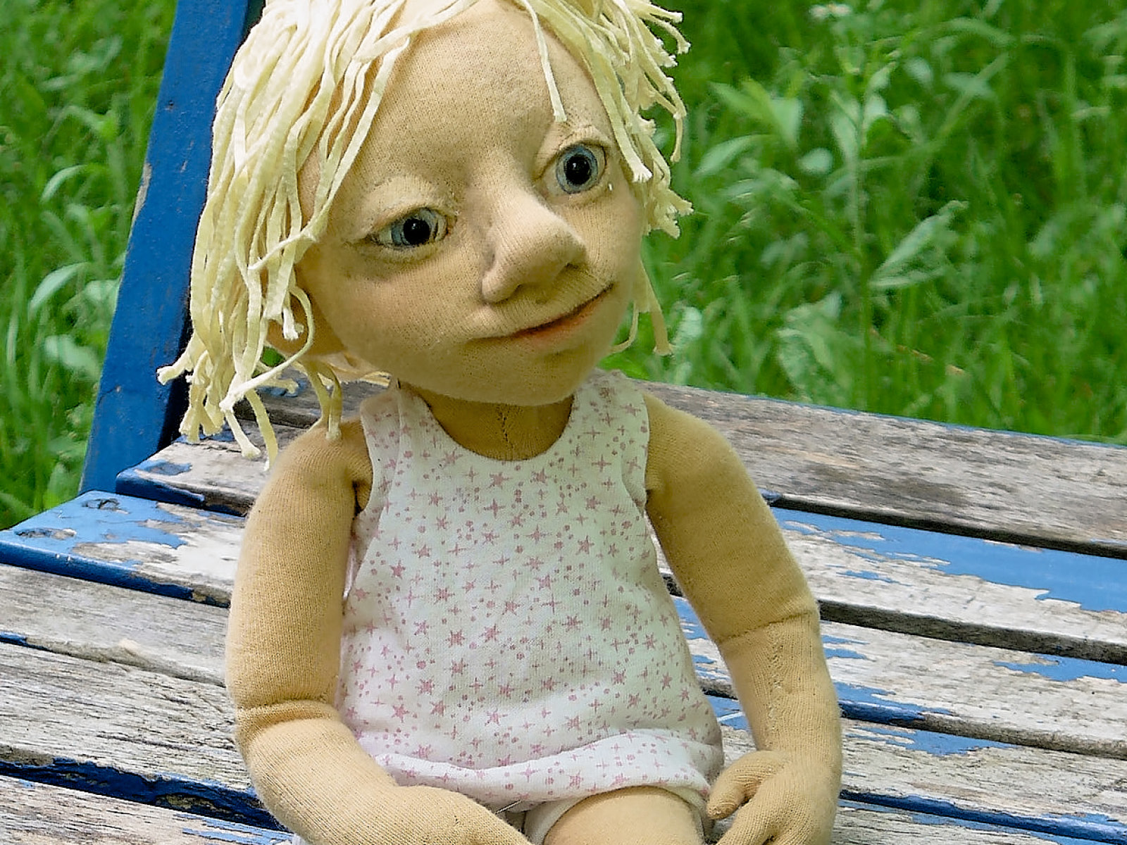 Die Puppe Lotta sitzt mit  Unterhemd und Unterhose, weißblonden strähnigen verwuschelten Haaren, großen hellblauen Augen und einem leicht lächelnden Mund auf einem blauen Stuhl in einer grünen echten Wiese.
